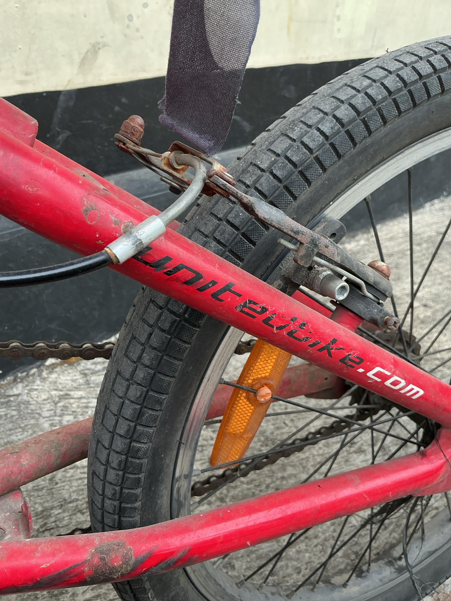 The image shows a close-up view of the rear wheel and frame of a red bicycle, likely in an outdoor urban setting in Magelang, Indonesia. The bicycle appears to be a BMX style, with thick tires and a chain visible. The frame has some rust and wear, suggesting it is an older or well-used bicycle. A portion of a dark fabric strap is visible at the top of the image. The text "unitebike.com" is visible in white lettering on the red frame. The background is out of focus, but appears to be a textured surface, possibly concrete or pavement. There are no people or animals present in the image.