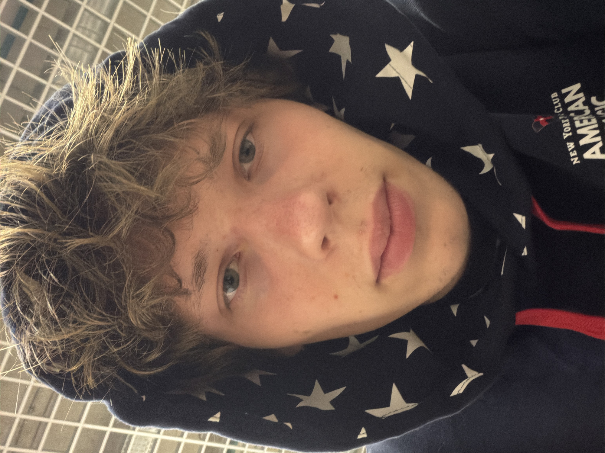 The image shows a close-up, selfie-style shot of a young Caucasian male with light brown, messy hair, wearing a navy blue hooded sweatshirt with white stars and red trim. The sweatshirt has some text on the lower right side that is partially visible, reading "NEW YORK CLUB AMERICAN MAGIC". The background appears to be an indoor setting with a tiled wall. There is no indication of the location being Nizhny Novgorod, Russia.