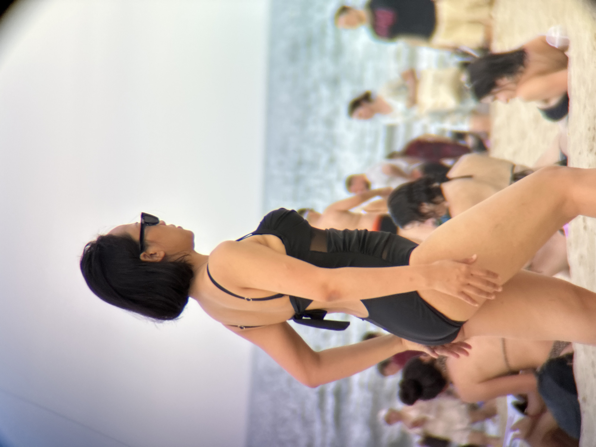 A woman in a black swimsuit with sheer mesh panels stands on a sandy beach, looking to her right. She has short dark hair and is wearing sunglasses. The ocean is visible behind her, and other people are blurred in the background. The scene appears to be a daytime shot on a beach, possibly in Da Nang, Vietnam, given the context provided.