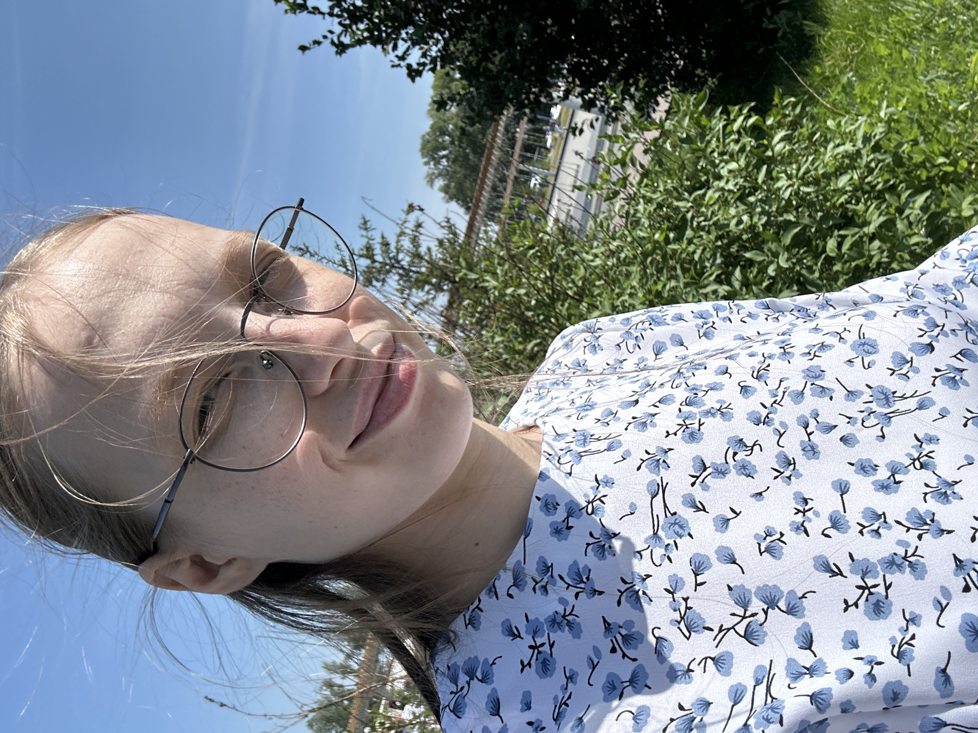A young woman with blonde hair and glasses is taking a selfie outdoors. She is wearing a white dress with a blue floral pattern. Her hair is blowing in the wind, partially obscuring her face. Behind her, there are green trees and bushes, and a bright blue sky. In the background, there is a structure with a wooden roof and some cars. The scene appears to be in a park or natural area within the city of Металлплощадка, Russia.