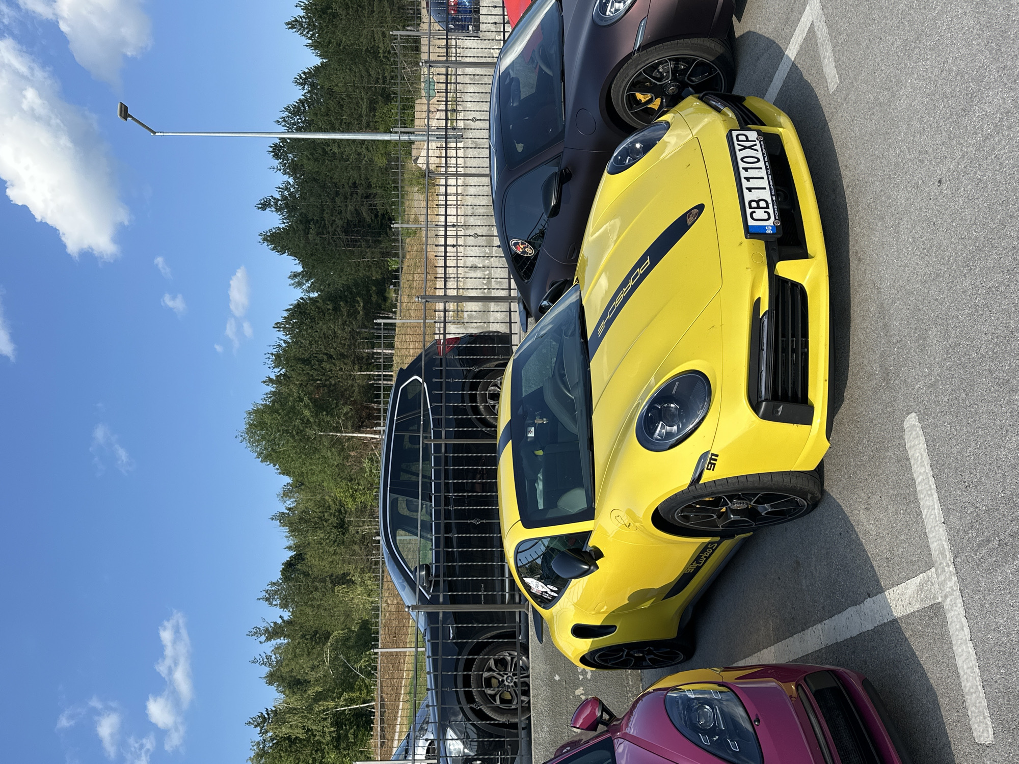 The image shows a parking lot in Kerenite, Bulgaria, with several high-performance cars. In the foreground, a bright yellow Porsche 911 Turbo S is parked, its front bumper visible with a Bulgarian license plate reading "CB 1110 XP". To its left, a portion of a dark pink car, possibly a Porsche, is visible. Behind the yellow Porsche, a dark brown Porsche is parked. Further back, a black SUV is parked behind a metal fence, and a line of trees forms the background. The sky is blue with scattered clouds.