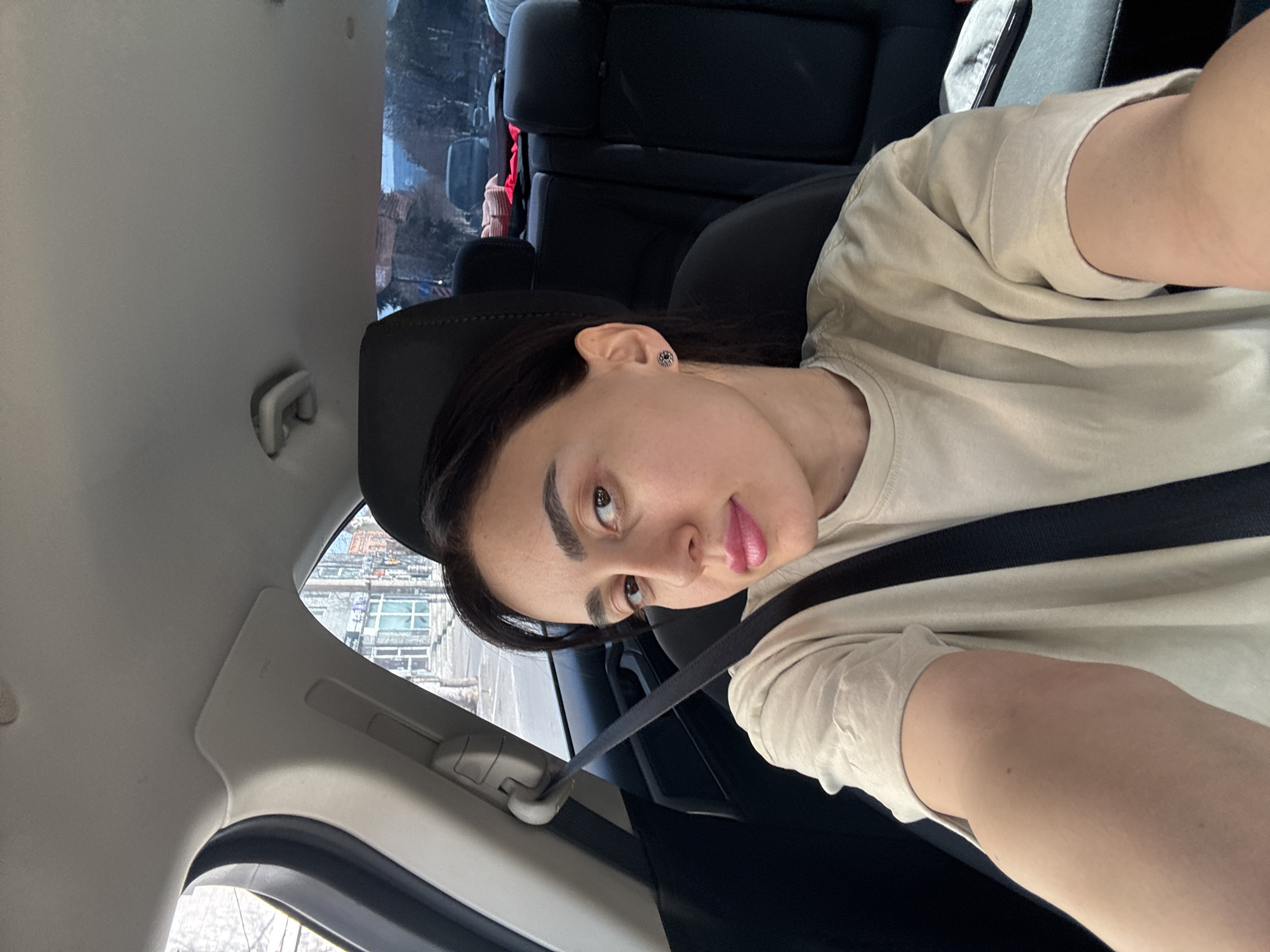 A person is seated in the driver's seat of a car wearing a seatbelt and a beige t-shirt. The car is likely in motion based on the blurred background. In the background, through the windows, there are buildings which appear to be located in an urban environment, perhaps the city of Tbilisi, Georgia. There are parked cars in the background. The person is looking at the camera with a neutral expression. Their hair is dark and pulled back from their face. The car's interior is predominantly black and grey.