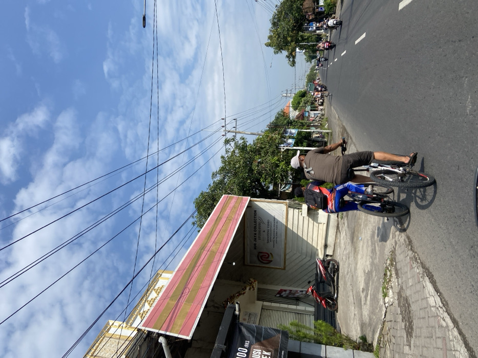 Two men are riding bicycles down a paved road in Kecamatan Tanggulangin, Indonesia, located at latitude: -7.497, longitude: 112.686. The man in front is wearing a brown t-shirt and a white cap. The man behind him is wearing a blue and red shirt and a black backpack. There are many power lines overhead and several buildings lining the road. The sky is mostly blue with some white clouds. Other vehicles can be seen in the distance, including a motorbike and a few cars. There are several small trees along the street and some greenery in the background. The men are riding in the same direction, and there are no other people visible. The road is relatively wide, with no obstacles present. The ground is paved with a light grey asphalt. The day is clear and sunny.