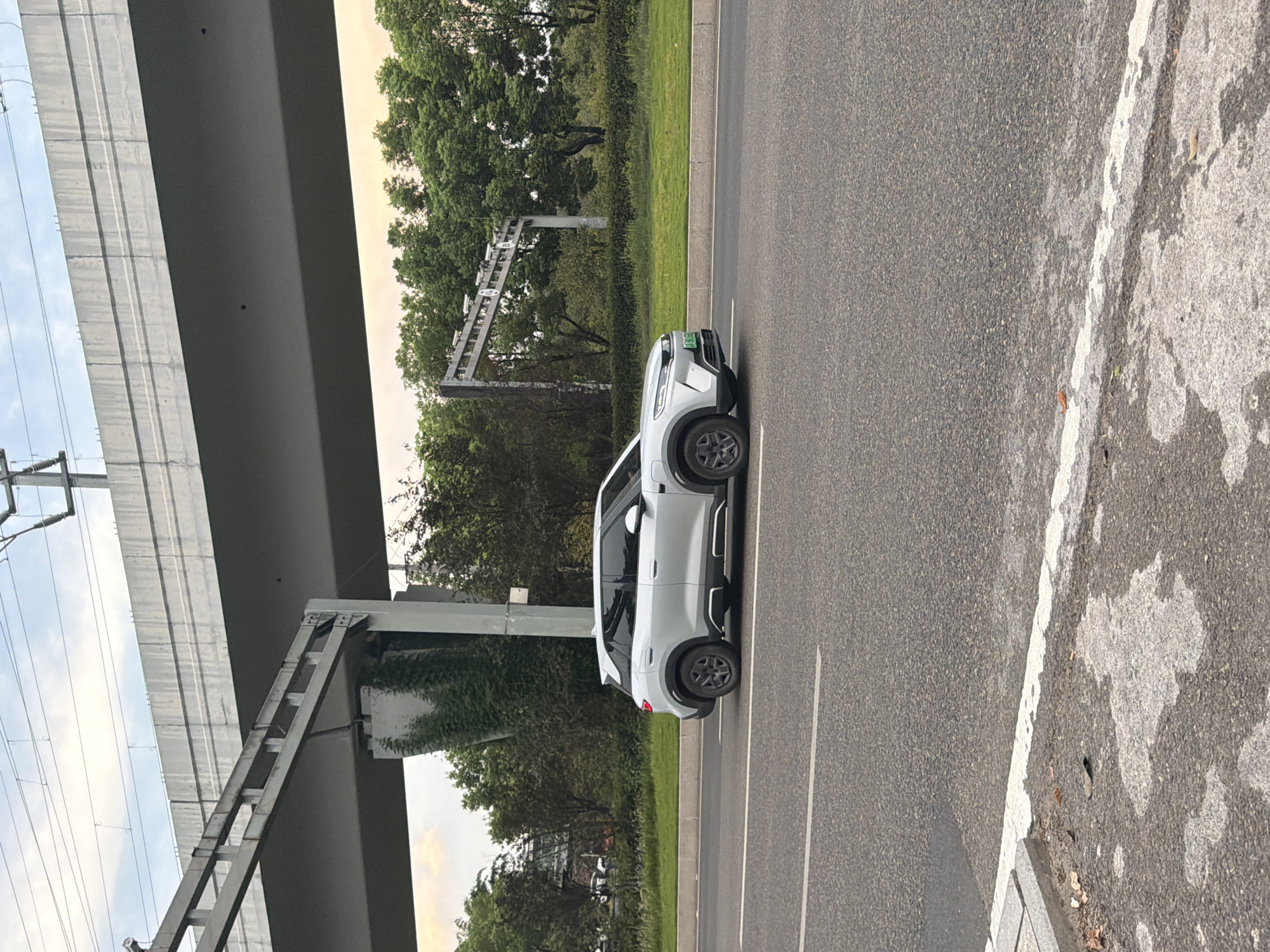 A silver SUV drives on a paved road in Jiangjin, China. The car is moving from left to right in the frame. The SUV has tinted windows and black rims. Above the car, a large concrete overpass with overhead electrical wires is visible. Trees and vegetation line the side of the road. A traffic camera gantry is also present in the background. The sky is overcast with hints of sunset colors. The road surface is asphalt with white lane markings. A concrete curb with some debris is present in the foreground.