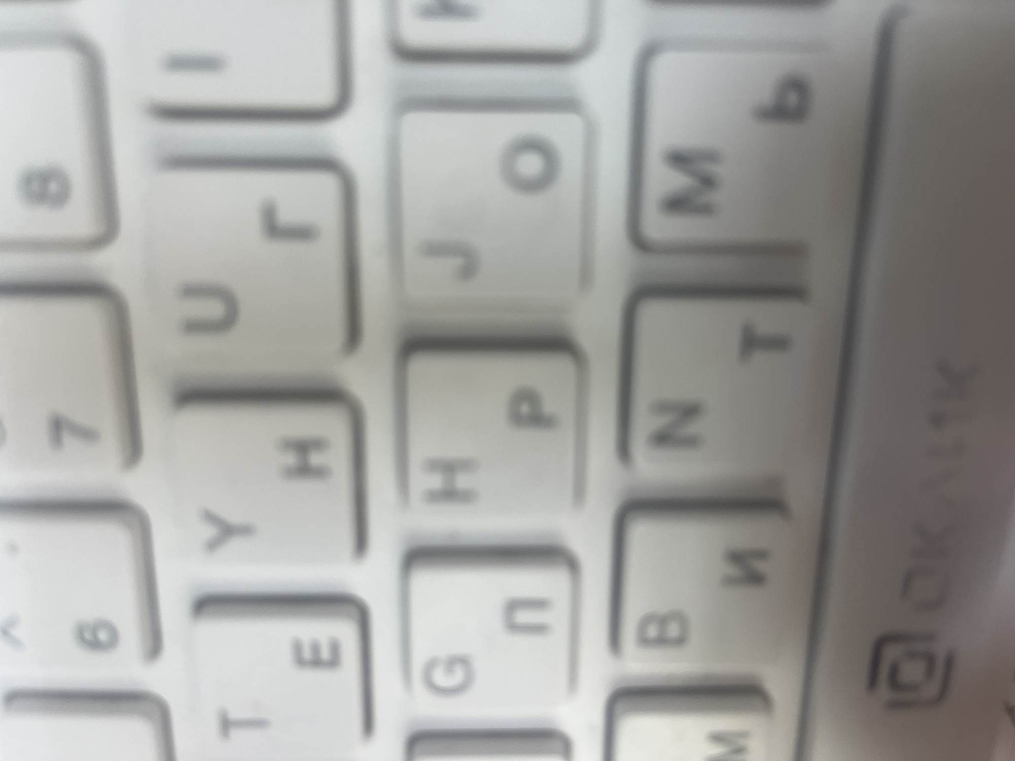 The image is a close-up, blurry photograph of a portion of a white keyboard. The keys are largely visible, showing various letters and numbers in Russian. Specifically, the keys visible include numbers 6, 7, and 8, and letters T, E, Y, H, U, R, I, G, П, H, P, J, O, M, B, N, M, and b. The keyboard appears to be from a laptop or a compact desktop computer. At the bottom of the image, a portion of a spacebar or a similar larger key is visible with the text "OK..." printed on it, along with an icon of a square with a smaller square inside it. The lighting suggests an indoor environment, and the overall quality of the image is low, with a lack of sharp focus. The image does not contain any elements that would place it in the city of Anapa, Russia, or any other specific geographic location.