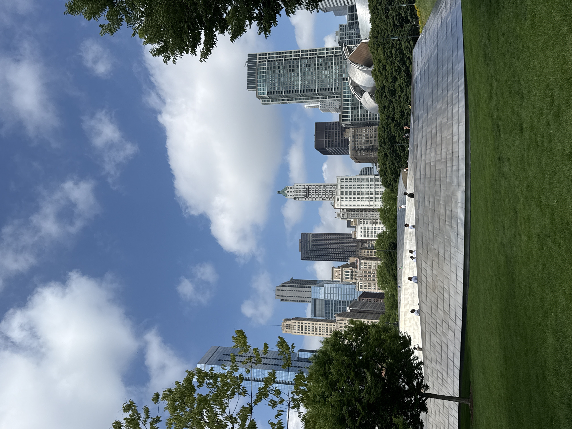 The image depicts a sunny day in Chicago, United States of America. The scene is a cityscape with skyscrapers visible in the background under a clear blue sky dotted with white clouds. In the foreground, there's a large, curved, polished metal structure with a reflective surface. Adjacent to this structure is a vibrant green lawn. Several people are seen on and around the metal structure, appearing to be enjoying the outdoors and interacting with the artwork. Trees with lush green leaves frame the scene, adding a touch of nature to the urban environment. The time of day appears to be midday, given the bright sunlight casting shadows. The overall atmosphere is one of leisure and appreciation for public art and green spaces within a bustling city.