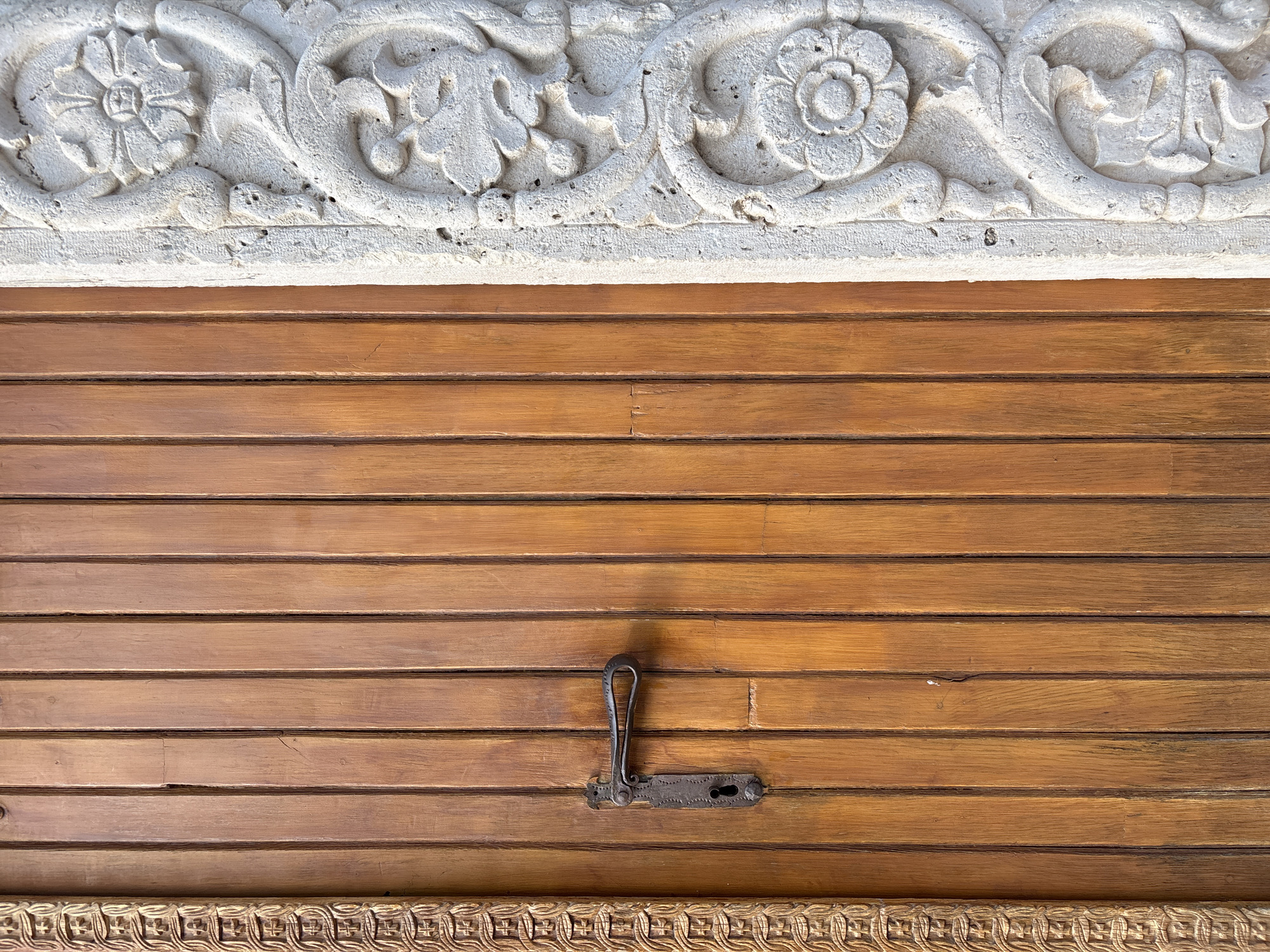The image shows a close-up of a wooden door with ornate carvings on the adjacent stone. The door is made of vertical wooden planks with a warm, reddish-brown hue. A dark metal door handle and lock mechanism are mounted on the door, positioned to the left of the center. To the right of the door, a stone pillar is visible, featuring intricate white carvings of floral and scrollwork motifs. The stone appears weathered and textured. The overall scene suggests an old architectural element, possibly part of a historic building in Budva, Montenegro, given the context. There are no people or animals in the image, and no discernible action is taking place. The lighting appears natural, casting subtle shadows that highlight the textures of the wood and stone.