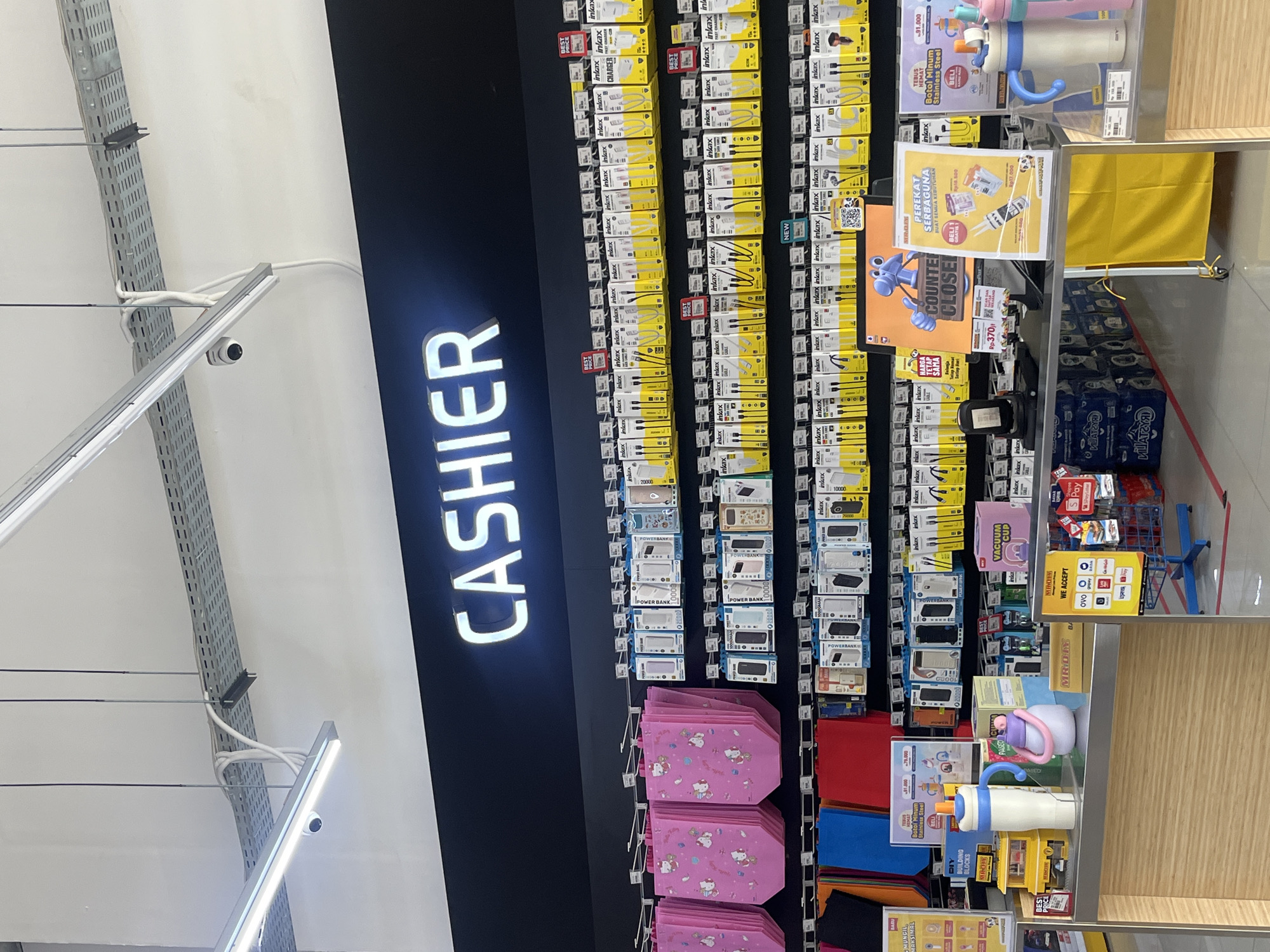 An indoor retail store, likely a discount or general merchandise shop, is depicted, specifically focusing on a cashier counter area. The image is taken at a tilted angle, showing overhead lighting and cable trays mounted on the white ceiling.
The most prominent feature is a brightly lit "CASHIER" sign displayed vertically on a dark wall. To its right, a wall display features numerous packaged electronic accessories, predominantly branded "inkax," including chargers, power banks, and cables, with some marked "BEST PRICE" and "NEW." Below these items, stacks of pink shopping bags adorned with Hello Kitty characters are visible.
The cashier counter itself holds various products and promotional materials. Several colorful water bottles or tumblers, some advertised as "Botol Minum Stainless Steel" and part of a "TEBUS HEMAT" (save by redeeming) promotion for Rp31.000, are on display. Other items include "DIY BUILDING BLOCKS" and what appears to be "COOKIE TIME" snacks. A sign indicating "COUNTER CLOSED" is prominently displayed with a cartoon character, alongside another sign for "PEREKAT SERBAGUNA" (all-purpose adhesive) offering a "BELI 1 GRATIS 1" (buy 1 get 1 free) deal at Rp18.500 or Rp17.000. A smaller sign shows "Rp370." Payment options like "OVO," "Gopay," and "Express" are listed on a "WE ACCEPT" sign. Under the counter, several blue crates, possibly containing bottled beverages like "PASEO," are stacked.
No people are visible, and no specific activity is taking place. The scene suggests a typical retail environment in West Jakarta, Indonesia, indicated by the currency (Rp) and some product names/promotions in Indonesian. A security camera is visible near the ceiling.