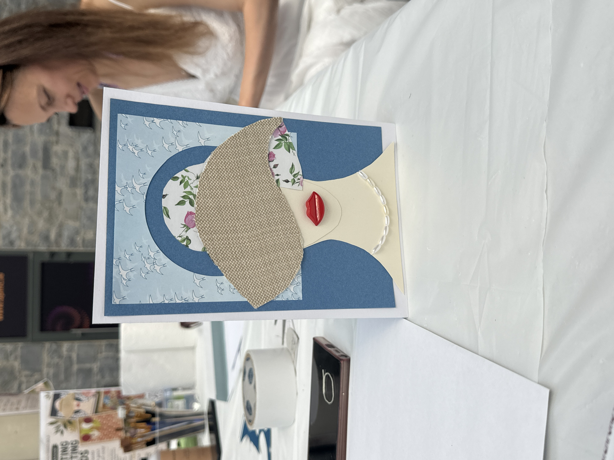 The image shows a handmade card featuring a stylized woman's face with red lips and a pearl necklace. The card is placed on a white tablecloth, with various crafting supplies in the background, including brushes, paper, and a roll of tape. A woman in a white dress is visible in the upper right corner of the frame, looking down at something out of frame. The scene appears to be an indoor setting, possibly a craft fair or workshop. The stone wall visible in the background is characteristic of many buildings in Kilkenny, Ireland.