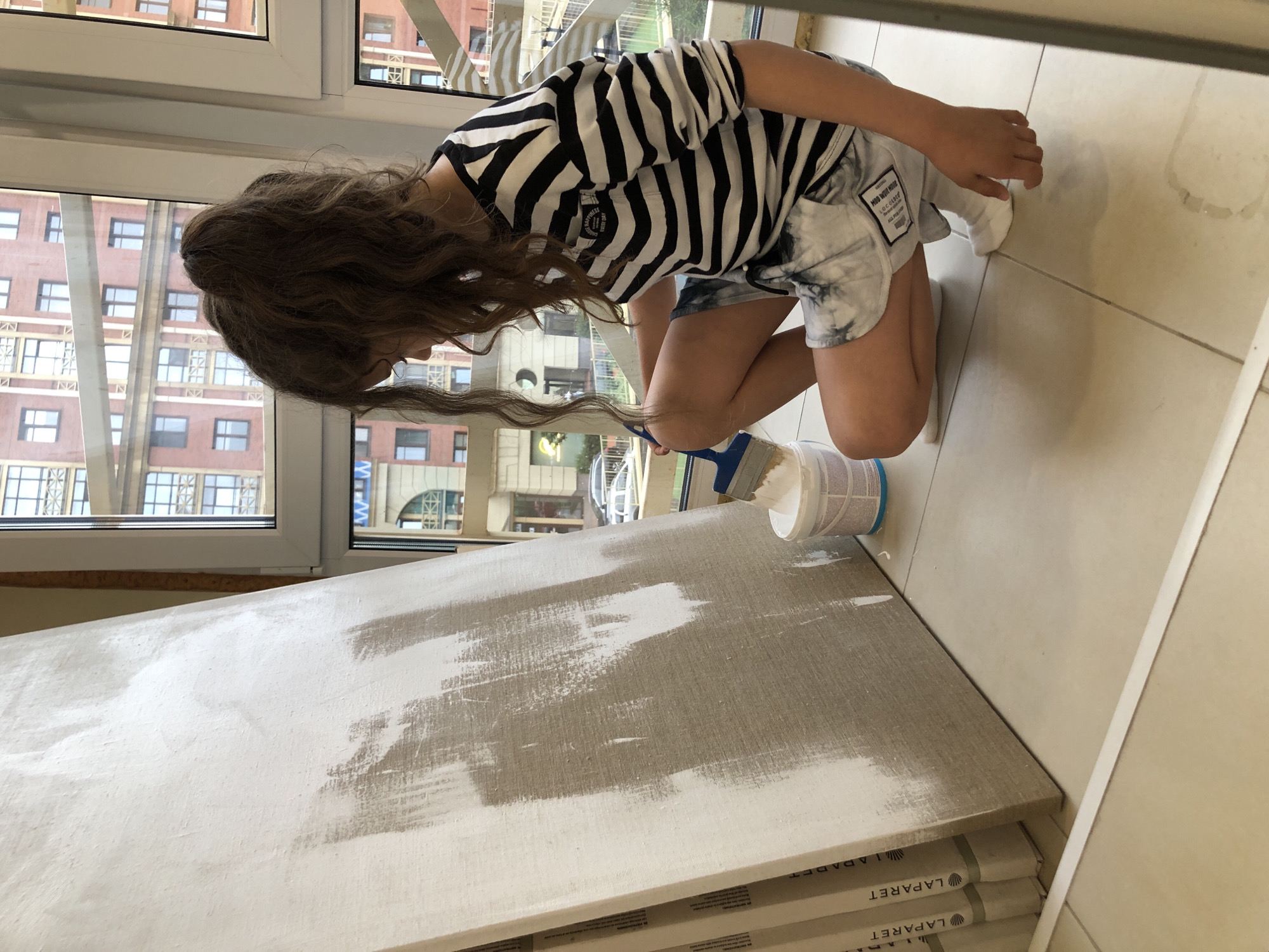 An unidentified person is squatting next to a large canvas that is partially covered in white paint. The canvas is leaning against a wall near a window. The person is wearing a black and white striped long-sleeved shirt and tie-dye shorts, with white socks and no shoes. They are holding a paintbrush that is dipped in white paint from a white bucket. The background shows a building with multiple windows, suggesting an urban environment. The floor is tiled. The person's face is not visible.