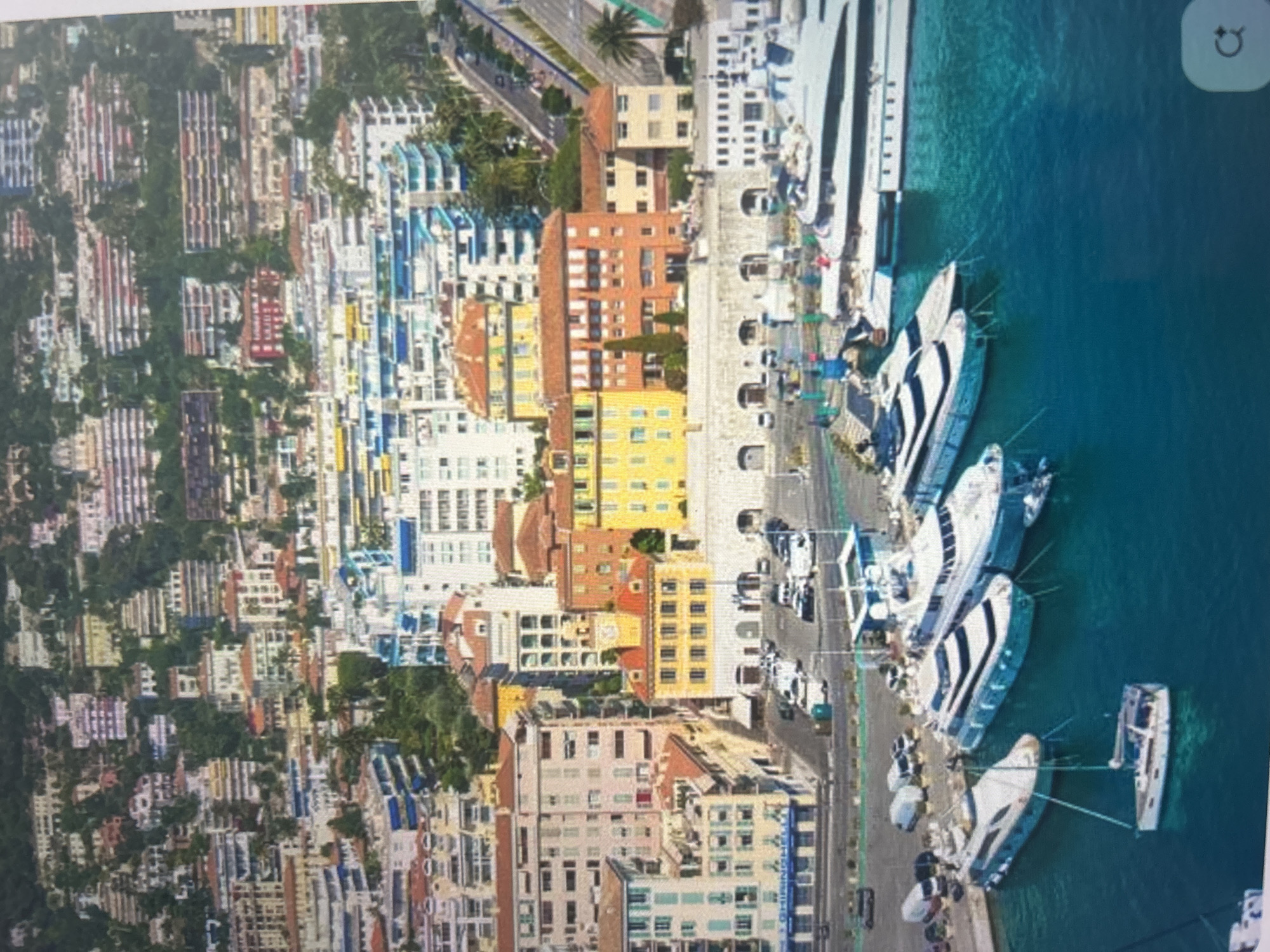 This aerial view captures a vibrant coastal city or town, likely in a Mediterranean region, with a prominent harbor. The scene is brightly lit under clear daytime skies.
On the land side, a dense array of multi-story buildings stretches across the landscape, displaying a rich palette of colors including yellows, oranges, reds, blues, and whites. These structures are interspersed with lush green trees and vegetation, suggesting a verdant environment. Roads wind through the urban area, with several cars visible, indicating active city life. Further up the hillside, more residential buildings line the slopes.
To the right, a large expanse of clear, turquoise water forms a bustling port or marina. Numerous boats and yachts of various sizes are docked along extensive quays and piers. Many of these appear to be large, luxurious yachts with sleek designs, while smaller vessels are also present. The water's clarity allows some underwater details to be visible near the docks.
A wide road runs parallel to the waterfront, separating the urban buildings from the harbor, and cars can be seen traveling along it. Palm trees are scattered throughout the scene, particularly near the waterfront and roads, enhancing the coastal atmosphere. In the top right corner, a white square icon with a circular arrow is visible, suggesting this is a screen capture. The overall impression is that of a picturesque and affluent coastal destination.