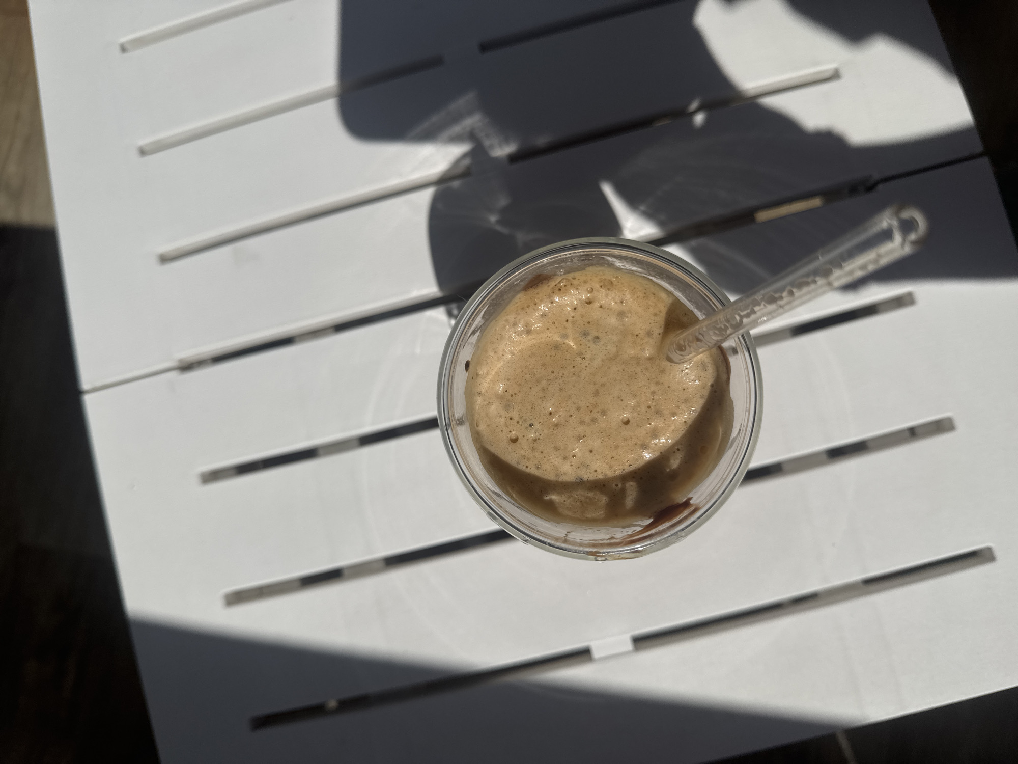 A top-down shot showcases a clear glass cup filled with a frothy, light brown beverage, likely coffee or a similar drink, topped with a foamy crema. A transparent plastic straw is inserted into the drink, with its textured surface visible. The cup rests on a white slatted table, its surface marked by distinct shadows cast from an unseen object, suggesting it's outdoors during the daytime. The shadows, sharp and elongated, imply the sun is at a moderate angle, possibly late morning or early afternoon. The slatted design of the table creates a linear pattern that contrasts with the roundness of the cup and the organic texture of the foam. The background shows the continuation of the white table and darker areas suggesting shade or the edge of a patio. The overall scene evokes a relaxed, casual setting, possibly a cafe or a personal outdoor space on a sunny day. The image's crisp focus on the drink and table suggests a clear day with ample sunlight. The location context of Varna, Bulgaria, adds a Mediterranean or Eastern European feel, where outdoor cafes and enjoying beverages in the sun are common. No visible text is present in the image.