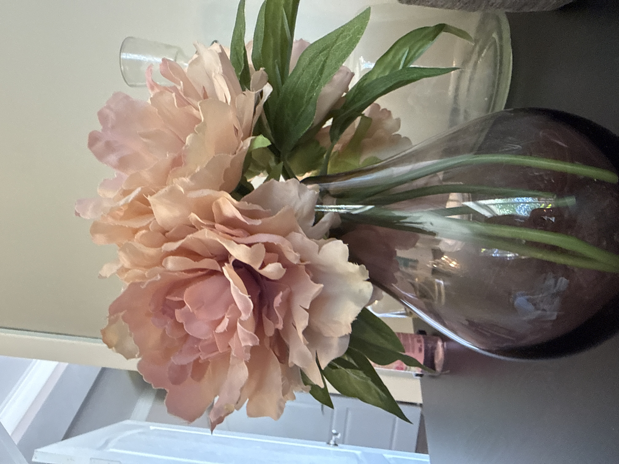 A close-up, eye-level indoor shot depicts a bouquet of three large, pale pink, peony-like artificial flowers with green leaves, arranged in a translucent, dark amber or smoky purple glass vase. The vase rests on a dark, matte-finished surface, possibly wood or laminate. Visible through the vase are multiple green stems of the flowers.
To the left of the main vase, a portion of a white door is visible, slightly ajar, with a metallic doorknob and white architectural molding above it. Behind the main vase, on the dark surface, a slender, light pink bottle or container is partially visible.
To the right and slightly behind the main vase, a clear glass object, possibly another vase or candlestick holder with a wide base, is present. Further to the right, a textured, light grey or off-white object is partially discernible. The background consists of a plain, light cream-colored wall. Reflections are visible on the surface of the dark vase, including a light blue rectangular reflection on its lower left and a brighter, irregular smudge on its front-right curve. The scene is illuminated by soft, diffuse lighting, casting minimal shadows. The location is in the city of Unknown, Unknown.