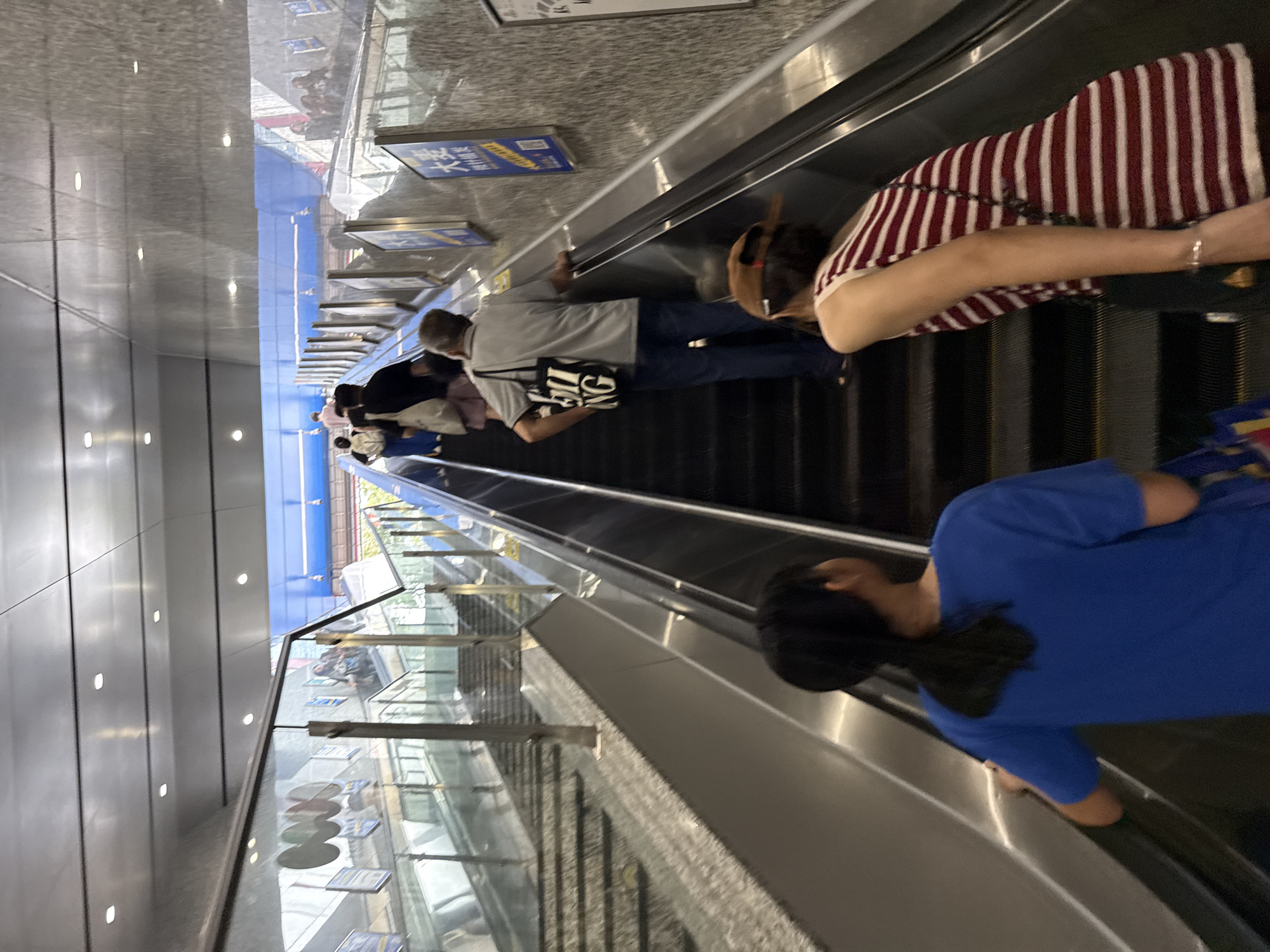 In Haizhu District, China, an outdoor escalator ascends towards a brightly lit area. A group of people are seen on the escalator. In the foreground, two individuals are ascending; one, a young person with dark hair pulled back in a ponytail, wears a blue t-shirt. Next to them, a person wearing a striped top and a baseball cap is also on the escalator. Further up the escalator, a man in a grey t-shirt and blue jeans carries a black tote bag with white lettering. Several other individuals are visible further up the escalator, some appearing to be in transit. The escalator is made of dark metal and is flanked by reflective glass barriers. The environment appears to be a transit hub or public space, with signage visible on the walls.