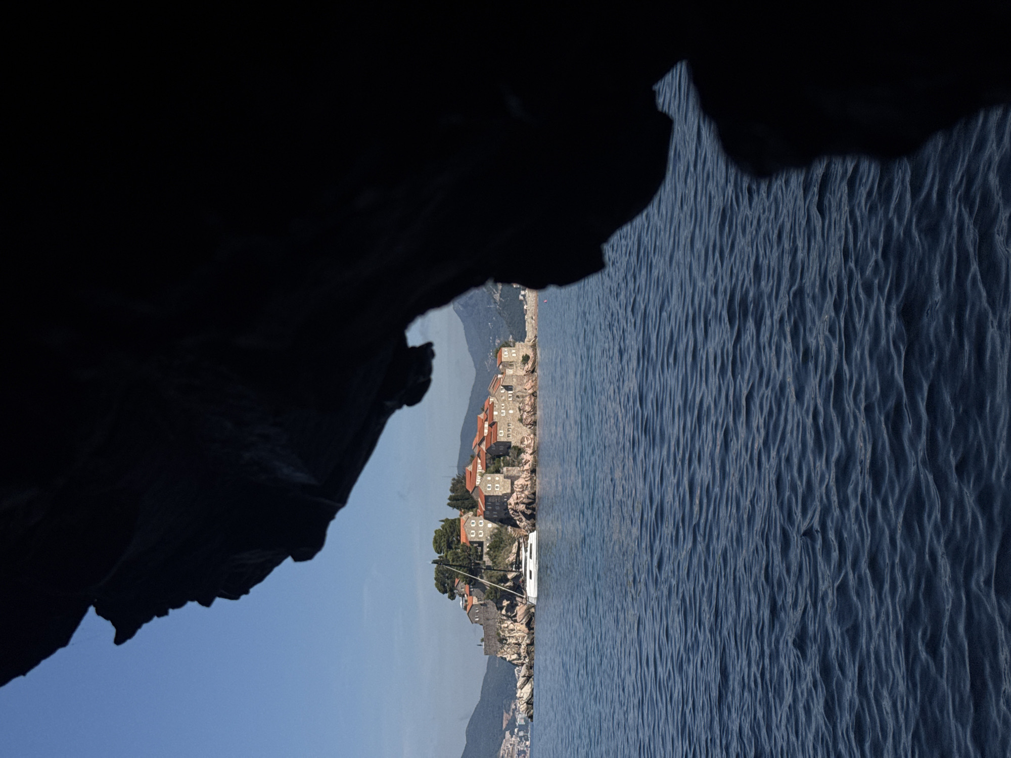 The image depicts a view of Sveti Stefan, a fortified island town in Montenegro, from a dark foreground element, likely a cave or overhang. The island is characterized by its cluster of stone buildings with orange tiled roofs, situated on a rocky outcrop. A white sailboat with a tall mast is moored in the calm, deep blue water in front of the island. The background shows rolling green hills and a clear blue sky. The overall scene suggests a serene coastal landscape.