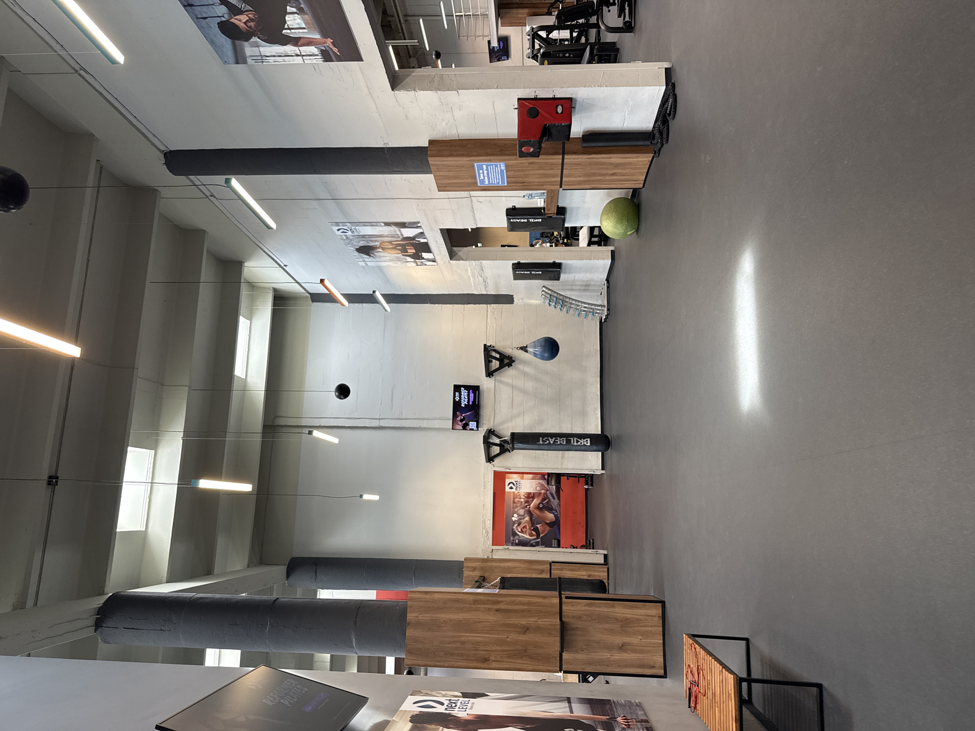 The image depicts the interior of a gym, likely located in Sofia, Bulgaria. The gym has a high ceiling with visible beams and linear light fixtures. The floor is gray and appears to be made of concrete or a similar material. Exercise equipment and training tools are placed throughout the space, including a boxing bag, weights, and machines. Various posters and signage with the name "BRL BEAST" and "NEXT LEVEL" adorn the walls, suggesting the name of the gym or fitness brand. No people are visible in the frame. The overall environment suggests a modern and well-equipped fitness facility.