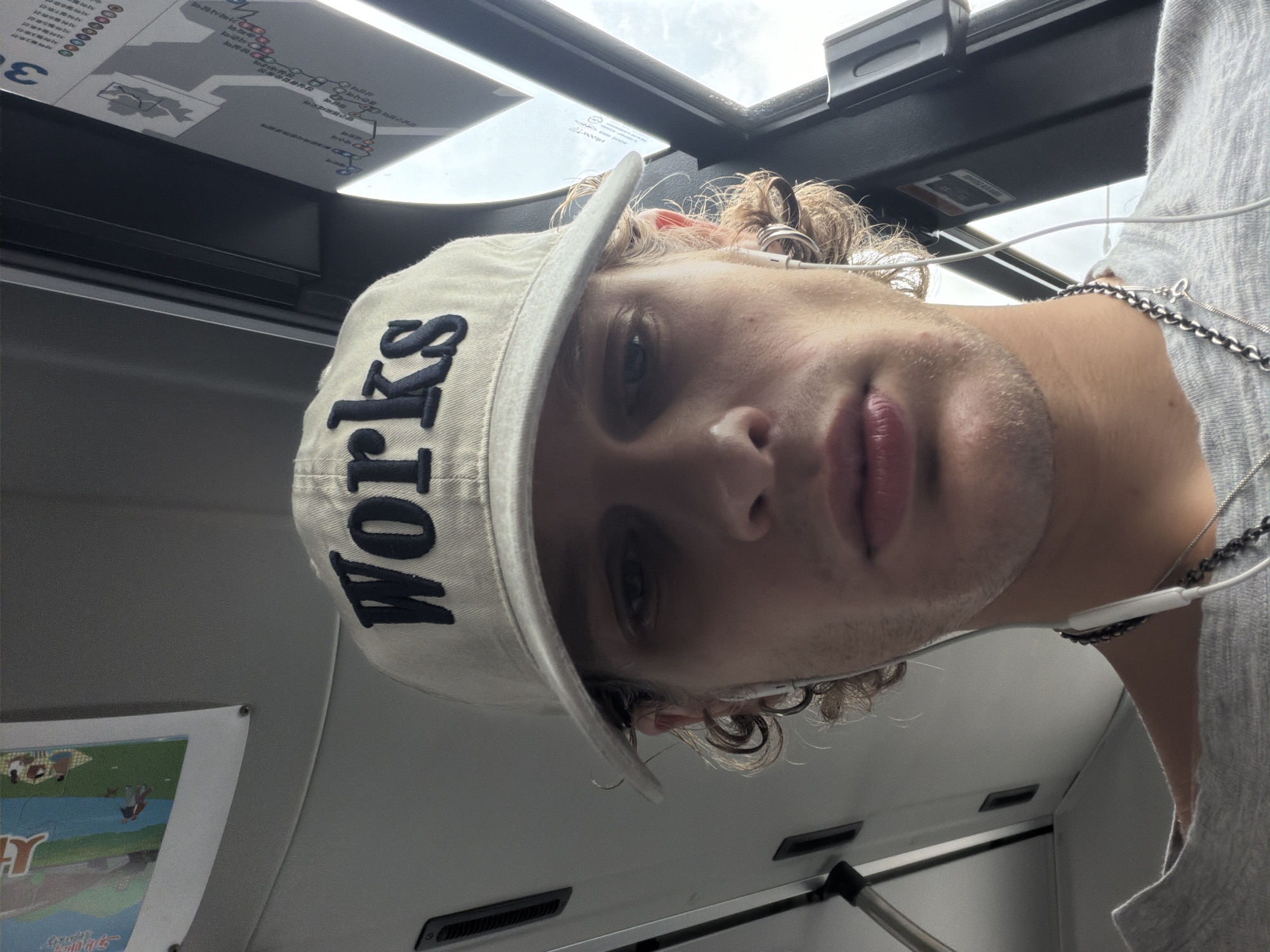 A close-up, low-angle selfie shows a young Caucasian person wearing a light-colored baseball cap with the word "Works" embroidered in dark blue lettering. The person has curly blonde hair peeking out from under the cap and is wearing white earbuds. They are also wearing a grey t-shirt and a silver chain necklace. The background appears to be the interior of a vehicle, possibly a bus or train, with grey paneling and a window. A map or schedule is visible on the wall to the right. The overall lighting suggests it is daytime. The image does not contain sufficient information to determine the location within Seoul, South Korea.