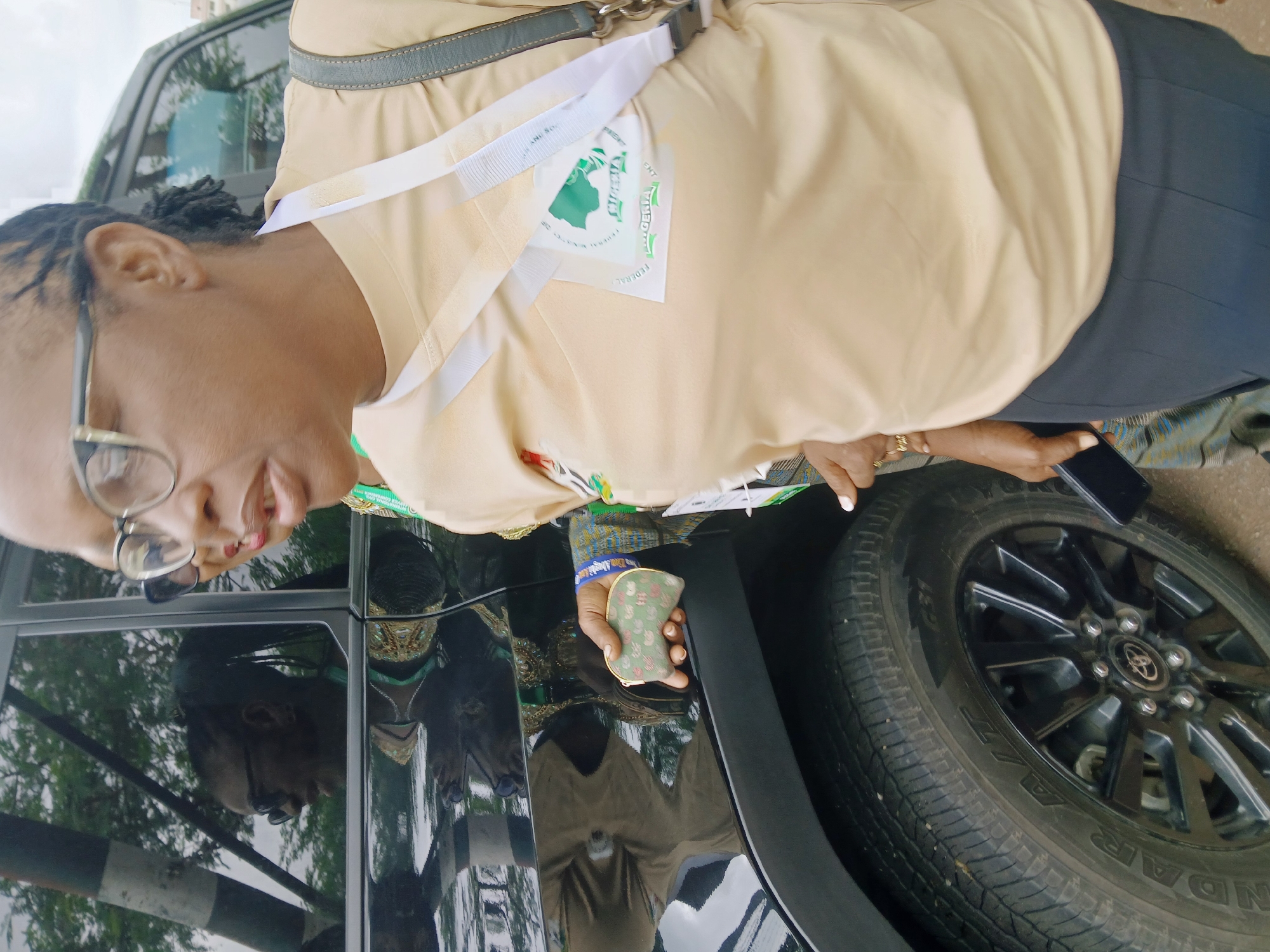A woman stands next to a black car, appearing to be a Toyota, likely within Abuja, Nigeria. She is wearing glasses, a light brown or beige shirt with a white lanyard displaying a green logo. The woman is holding a small green wallet with a floral pattern in her left hand and a black mobile phone in her right. She is wearing a black skirt. Her reflection is visible in the car's window.