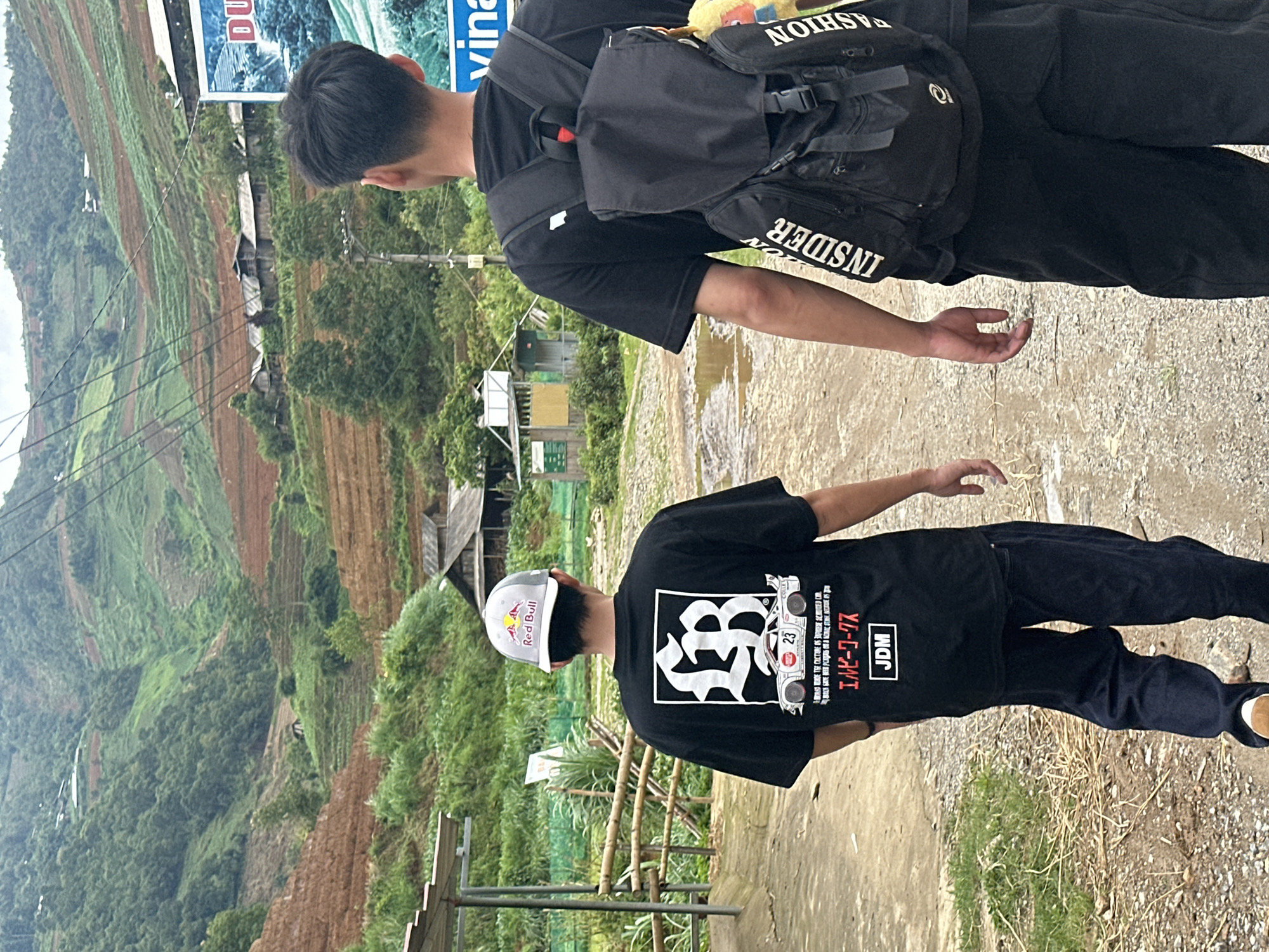 Two males are depicted walking away from the camera on an unpaved path in the mountainous region of Mù Cang Chải, Vietnam. The individual on the right, in the immediate foreground, wears a black short-sleeved t-shirt and a black backpack with "INSIDER" vertically printed on the left strap and "FASHION" vertically printed on the right strap. The second individual, positioned slightly to the left and further back, wears a grey and white Red Bull baseball cap, a black short-sleeved t-shirt featuring a large white graphic of an "LB" emblem, a white racing car with the number "23", Japanese text ("エルビーワークス"), and "JDM", along with dark blue trousers and visible brown shoes.
The path is dirt, interspersed with patches of grass and several puddles. To the left, a simple shelter structure with a corrugated roof and wooden posts is visible, alongside a bamboo fence. To the right, a partial blue and white billboard depicting a waterfall and text, including "DU" and "vina", is present, accompanied by a utility pole and numerous overhead power lines extending across the scene. The background is dominated by steep, rolling hills covered in dense green foliage and an extensive system of agricultural terraces. Some terraces are vibrant green with crops, while others appear reddish-brown, indicating bare soil or recent cultivation. Multiple small, rustic wooden houses with dark roofs are scattered among the terraced fields on the hillsides. The sky overhead is overcast.