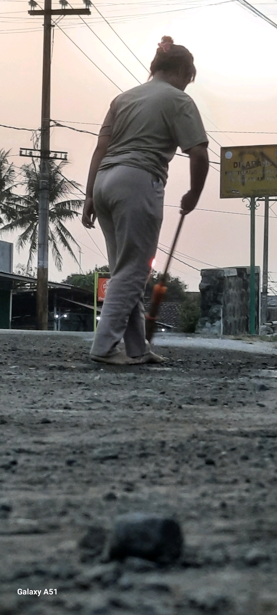 An outdoor, full-body, slightly low-angle shot shows a person sweeping the ground in Selopuro, Indonesia, against a backdrop of a power pole and palm trees. The person, from behind, is wearing a green t-shirt and light-colored pants and has their hair pulled up with a pink accessory. They are using a broom with an orange handle to sweep the dusty, gravelly ground. In the background, utility poles with electrical wires are visible, along with palm trees and a sign that reads "DILARANG MEROKOK" (Do Not Smoke). A partially visible building with lights and a stone wall are also present. The sky is overcast with a pale orange hue, suggesting dusk. In the foreground, a blurry, dark, rectangular object lies on the ground. The bottom of the image displays "Galaxy A51," indicating the phone model used to capture the image.