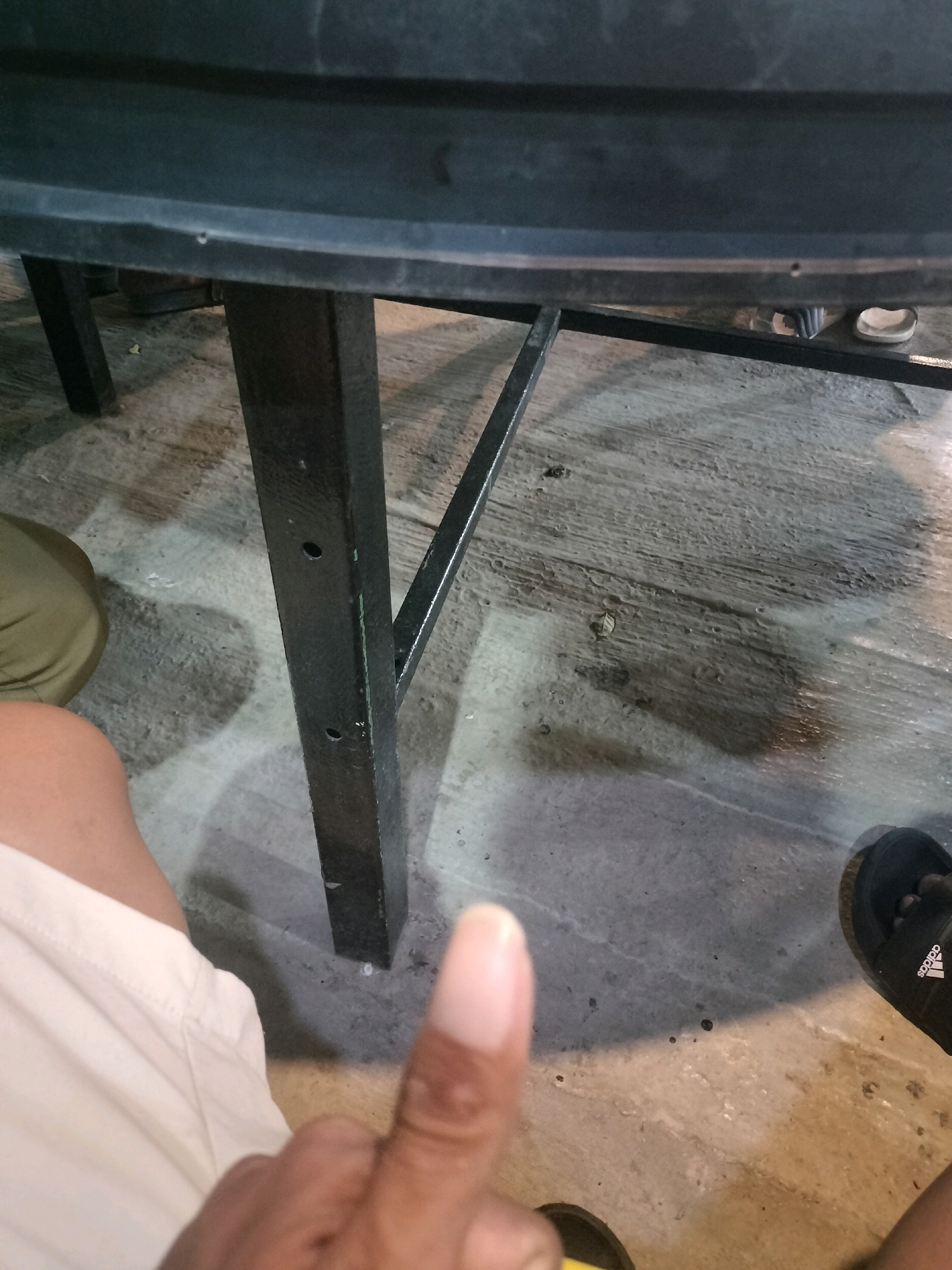 This is a low-angle, close-up image of a person's thumb and forearm, as well as a black table leg and the floor beneath it. The person is wearing light-colored shorts and a sandal with the Adidas logo is visible on their right foot. The floor appears to be concrete and is slightly wet in places. The table leg is made of dark metal and has visible screw holes. The scene appears to be outdoors in a public space, possibly a restaurant or cafe, in Batam, Indonesia. The lighting suggests it is daytime, but there are shadows cast by the table.