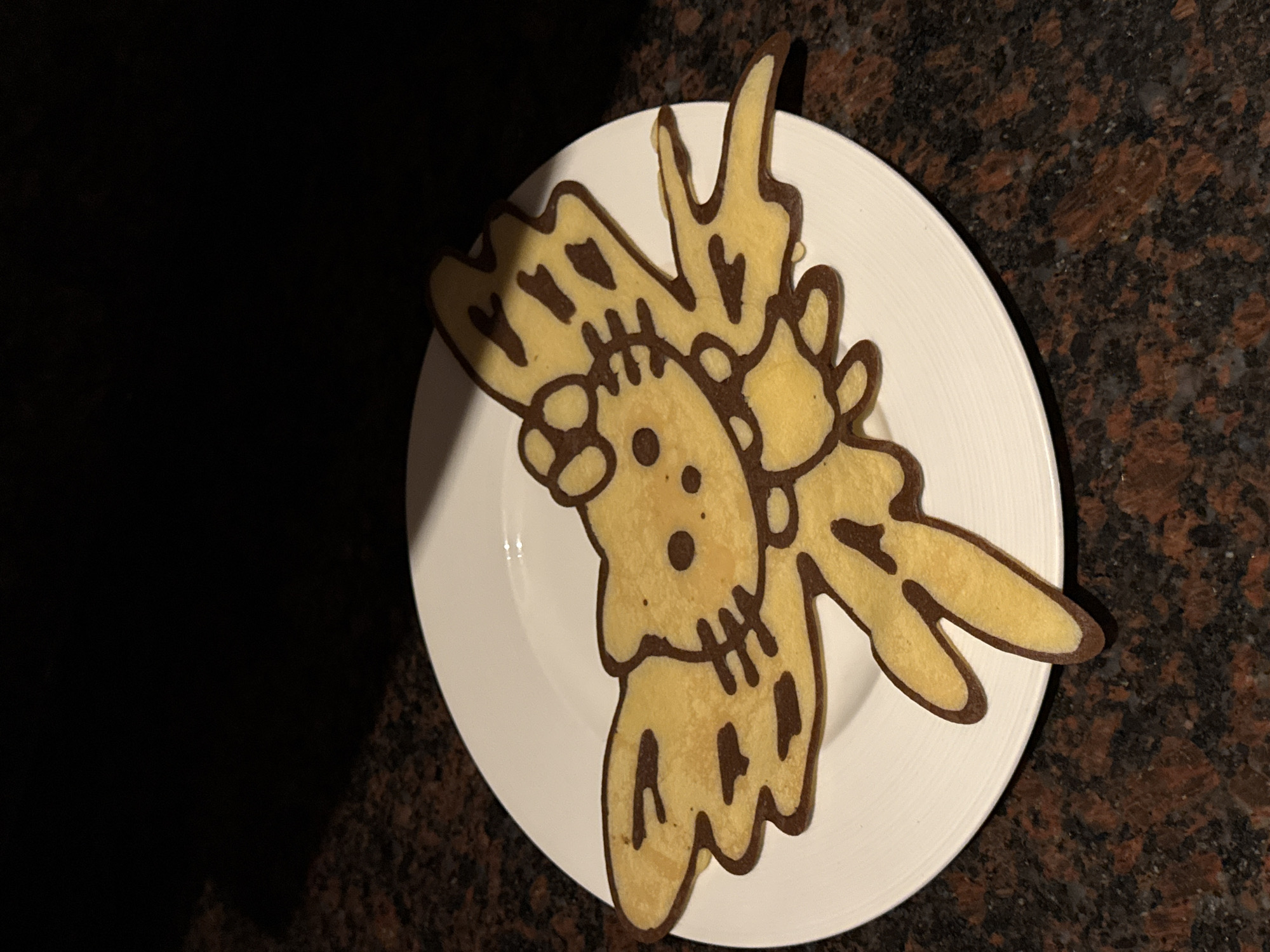 A Hello Kitty shaped pancake with dark brown outlines is presented on a white plate. The pancake has wing-like appendages and decorative spots on the wings. The plate is set on a speckled brown countertop. The background is dark and out of focus. The image was taken in Kuta Selatan, Indonesia.