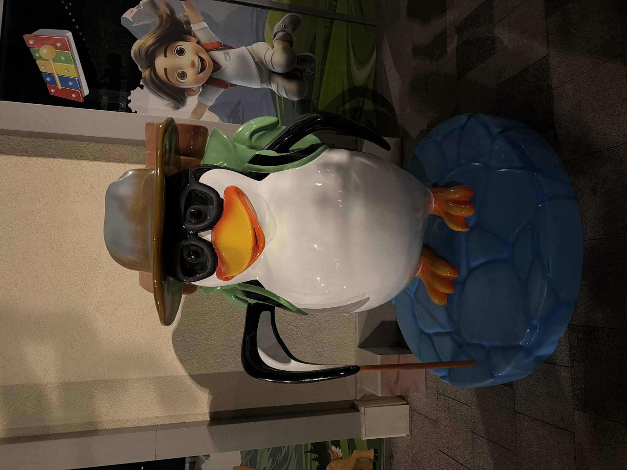 A statue of a penguin dressed as a hiker is displayed outdoors. The penguin is wearing a wide-brimmed hat and sunglasses, with a green backpack on its back. It stands on a blue, rounded platform that resembles ice. The penguin holds a walking stick in its left flipper. In the background, there are advertisements featuring animated characters and mountains, suggesting a theme park or recreational area. The ground is paved with interlocking stones. The lighting suggests it is evening or night. The image does not contain information pertaining to the city of Chinarkent, Uzbekistan.