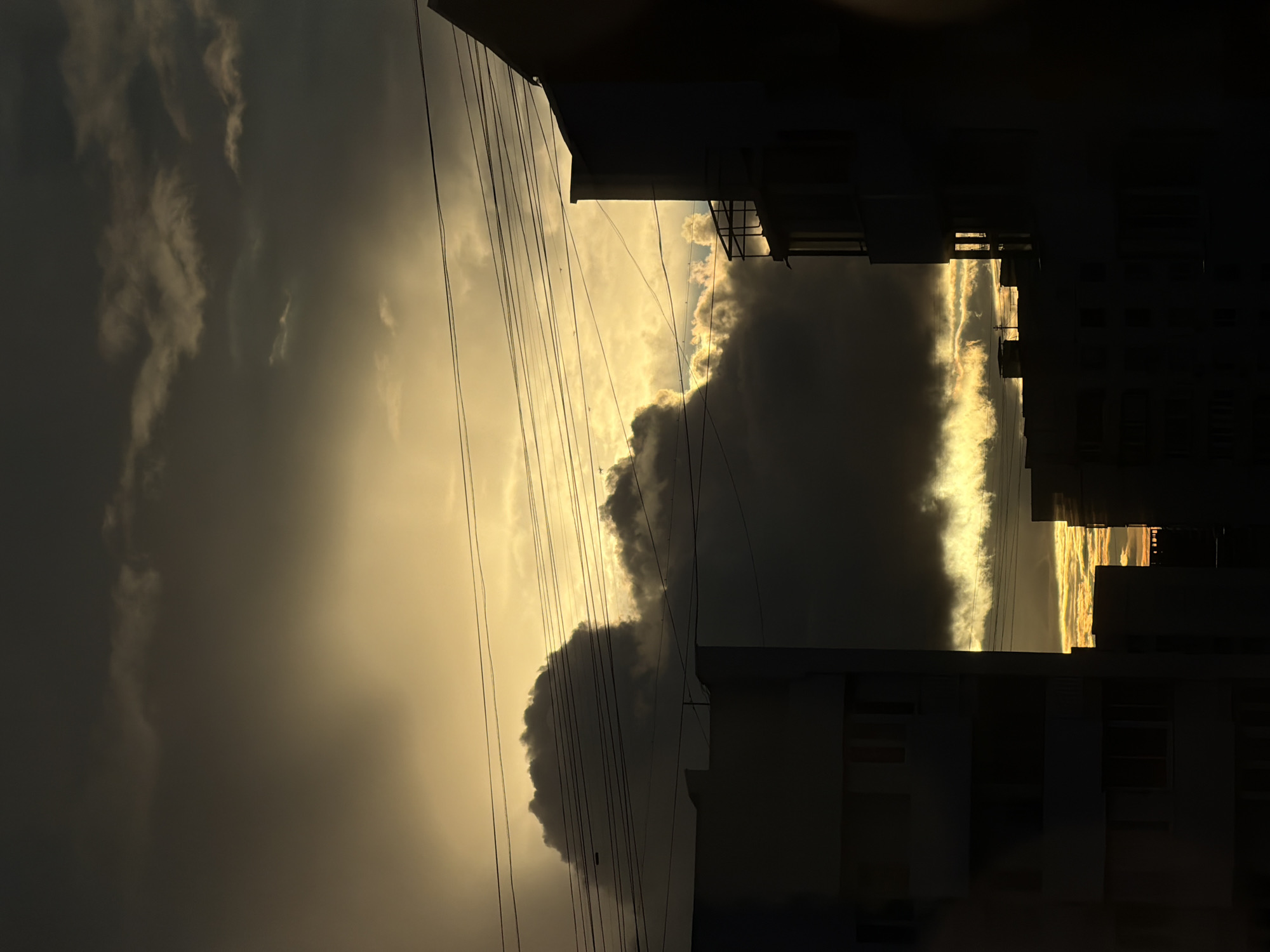 The image captures an obscured sunset in what appears to be an urban setting. The horizon is not visible. Dark, billowing clouds dominate much of the sky, obscuring the setting sun, but narrow slivers of golden light filter through, creating striking contrast. Power lines run across the view. Dark buildings, possibly apartments, are visible on the right. Based on the location provided (Khimki, Russia), the buildings and overall urban landscape suggest this setting. No people are visible.