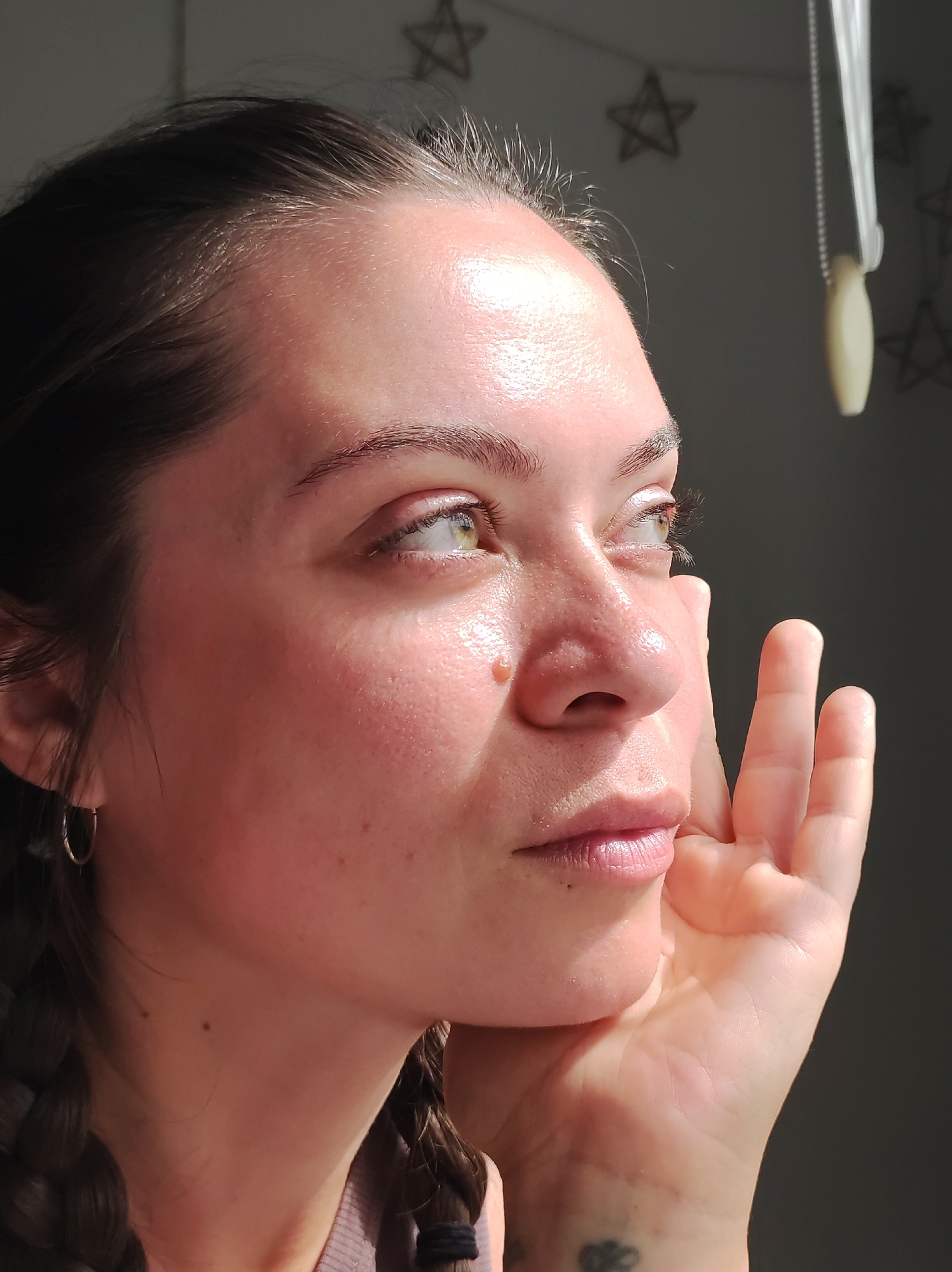 The photograph captures a close-up, side-profile of a young woman's face, illuminated by natural light filtering in from the right. Her eyes, a striking green, are looking upwards and to the right, catching the light. Her skin appears dewy and reflective, with visible pores and a slight rosy flush on her cheeks. A small mole is visible on her cheekbone, and her lips are slightly parted. Her hand is raised to her face, with fingers gently touching her cheek and chin. Her dark hair is pulled back, with one braid visible to the left and another towards the bottom of the frame. In the background, a dark wall is adorned with several star-shaped ornaments, and a light-colored pull cord hangs from the top right. The scene suggests an indoor setting, possibly a room with a window providing the directional light. There is no visible indication of the city of Chaikovsky, Russia.