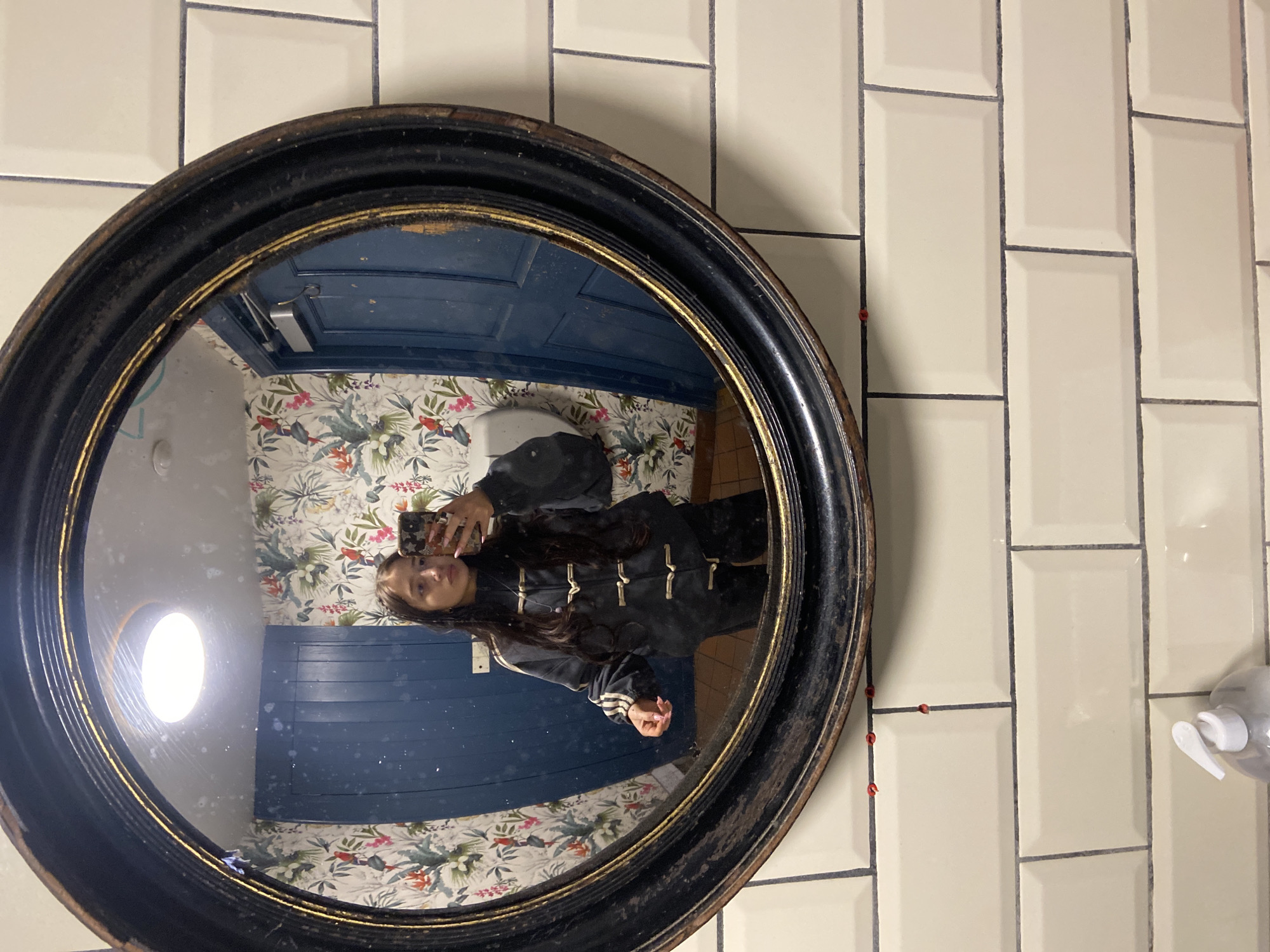 A female is reflected in a circular mirror. The mirror is framed with a black and gold ornate design and hangs against a wall tiled with rectangular, off-white tiles. The woman is holding a phone, presumably taking the photograph. Her hair is dark, and she is wearing a black jacket with gold embellishments. The reflection shows a bathroom with blue painted paneling and floral wallpaper. A toilet is visible in the background. Small red objects are affixed to the wall tiles around the mirror. The location is not identifiable as London, United Kingdom based on this image.