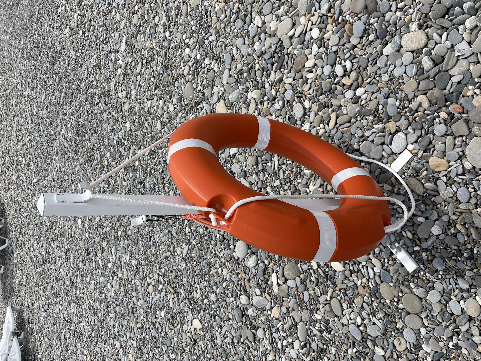 A bright orange life preserver with white stripes is attached by a rope to a white pole, which is mounted on a white metal stand. The life preserver is positioned in the center of the frame, against a backdrop of a pebble beach. The pebbles are of various sizes and colors, predominantly gray, brown, and white. In the upper left portion of the image, several empty white lounge chairs are visible on the beach, suggesting a recreational area. The scene appears to be on a beach in Sochi, Russia, likely a public area designated for swimming or water activities. No people are present in the image. The lighting suggests it is daytime, possibly overcast, with soft shadows.