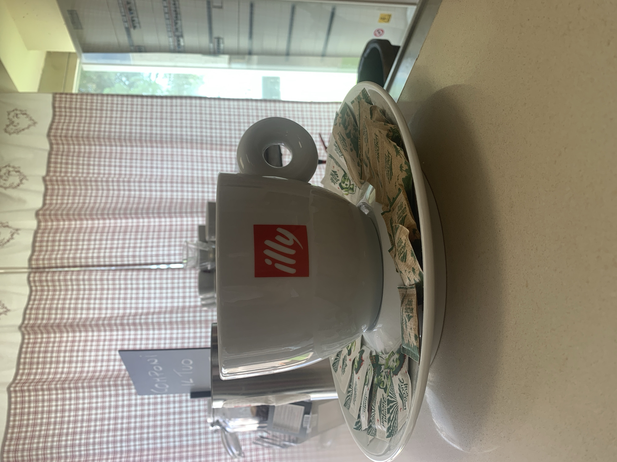 This is a medium shot, eye-level photograph taken indoors at what appears to be a cafe or restaurant in Foiano della Chiana, Italy. The primary subject is a white Illy coffee cup and saucer, positioned slightly off-center to the right. The cup has the distinctive red Illy logo on its side and a unique, round handle. The saucer is laden with several individually wrapped sugar packets, some appearing to be fruit-flavored.

In the background, to the left of the cup, is a stainless steel container, possibly a sugar dispenser or a grinder, and a glass jar with a lid. Behind these objects, a checkered curtain with a red and white pattern hangs, adorned with embroidered hearts. A small, dark sign with white lettering that reads "COMPONI IL TUO" (meaning "Compose yours" in Italian) is also visible near the curtain.

To the right of the cup, a window is visible, offering a glimpse of the outdoors, with green foliage suggested. The overall environment seems to be a clean, well-lit space, likely a counter area where patrons can prepare their coffee. The focus is sharp on the cup and saucer, with the background elements softened. The lighting appears to be natural, coming from the window, creating a slight shadow on the countertop. There are no people visible in the frame.