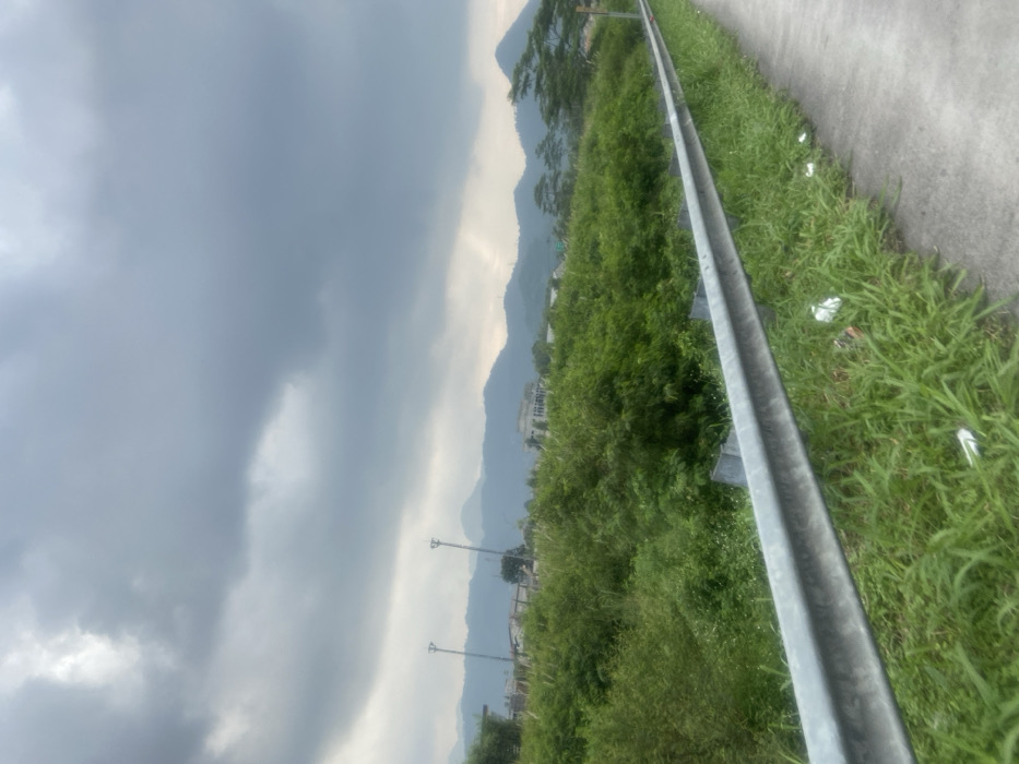 The image shows a view from a roadside in Margaasih, Indonesia, with a metal guardrail along a paved road. There is a dense growth of green foliage, shrubs and trees, in the foreground, and a row of hills in the distance. The sky is overcast with gray clouds. There are two tall, thin poles, possibly light posts, visible in the background. The image appears to have been taken on a cloudy, overcast day. The road is empty of traffic. The scene is typical of rural Indonesia, with the dense greenery and the rolling hills in the background.