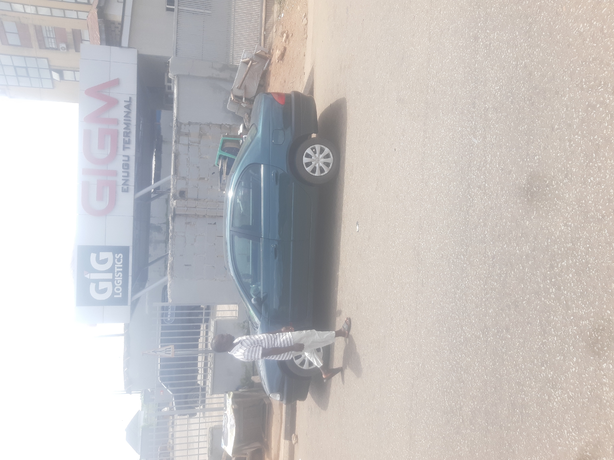 A man in a striped shirt and white pants walks on the right side of a dark green sedan. The car is parked parallel to a sidewalk in front of a building with a large sign that reads "GIG LOGISTICS" and "GIGM ENUGU TERMINAL". The sky is bright and overcast. The scene appears to be in Enugu, Nigeria.
