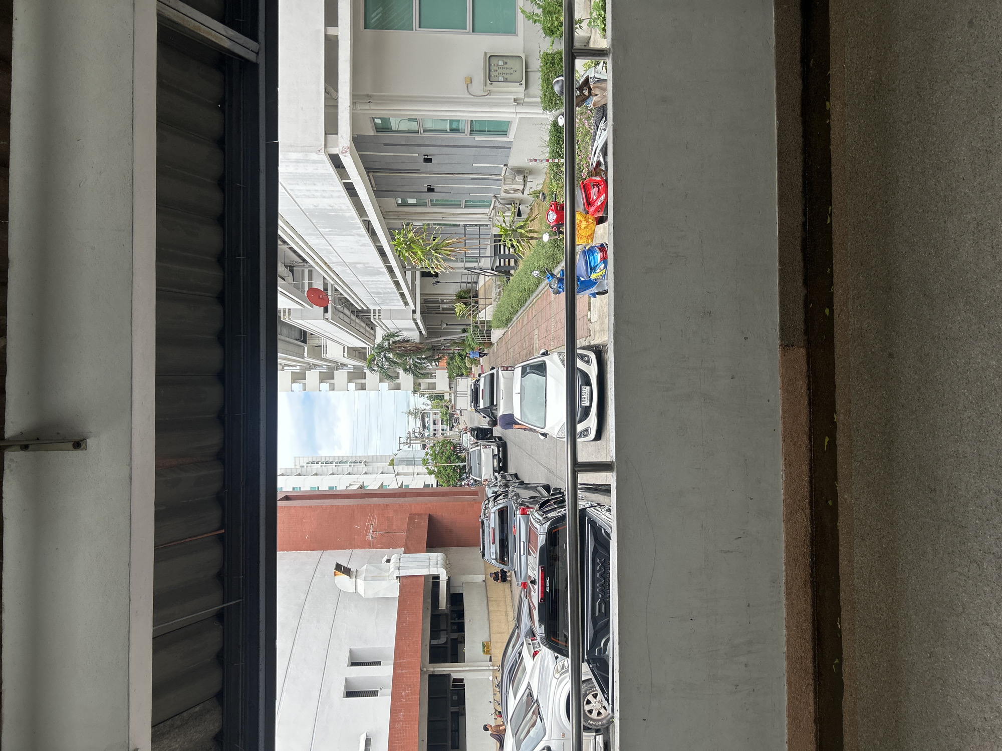 The image depicts a street scene in Tha Khlong Town Municipality, Thailand, viewed from an elevated position, possibly a walkway or balcony. In the foreground, a concrete railing separates the viewer from a paved area. Beyond the railing, a row of parked vehicles, including several pickup trucks and cars, lines the street. Some of these vehicles are silver, black, and white. A white sedan with a Thai license plate is visible on the right side of the frame.
Further down the street, more vehicles are parked, including white pickup trucks. A person in a blue shirt is walking on the left side of the street. On the right side of the street, a row of buildings is visible, characterized by white facades and multiple stories, some with balconies. Greenery, including bushes and trees, is present along the buildings and sidewalk. Several motorcycles are parked on the right side of the street near the buildings.
The architecture of the buildings suggests a modern urban environment. The sky is partly cloudy. The overall impression is of a typical day in a Thai urban area, with parked vehicles and pedestrian activity.