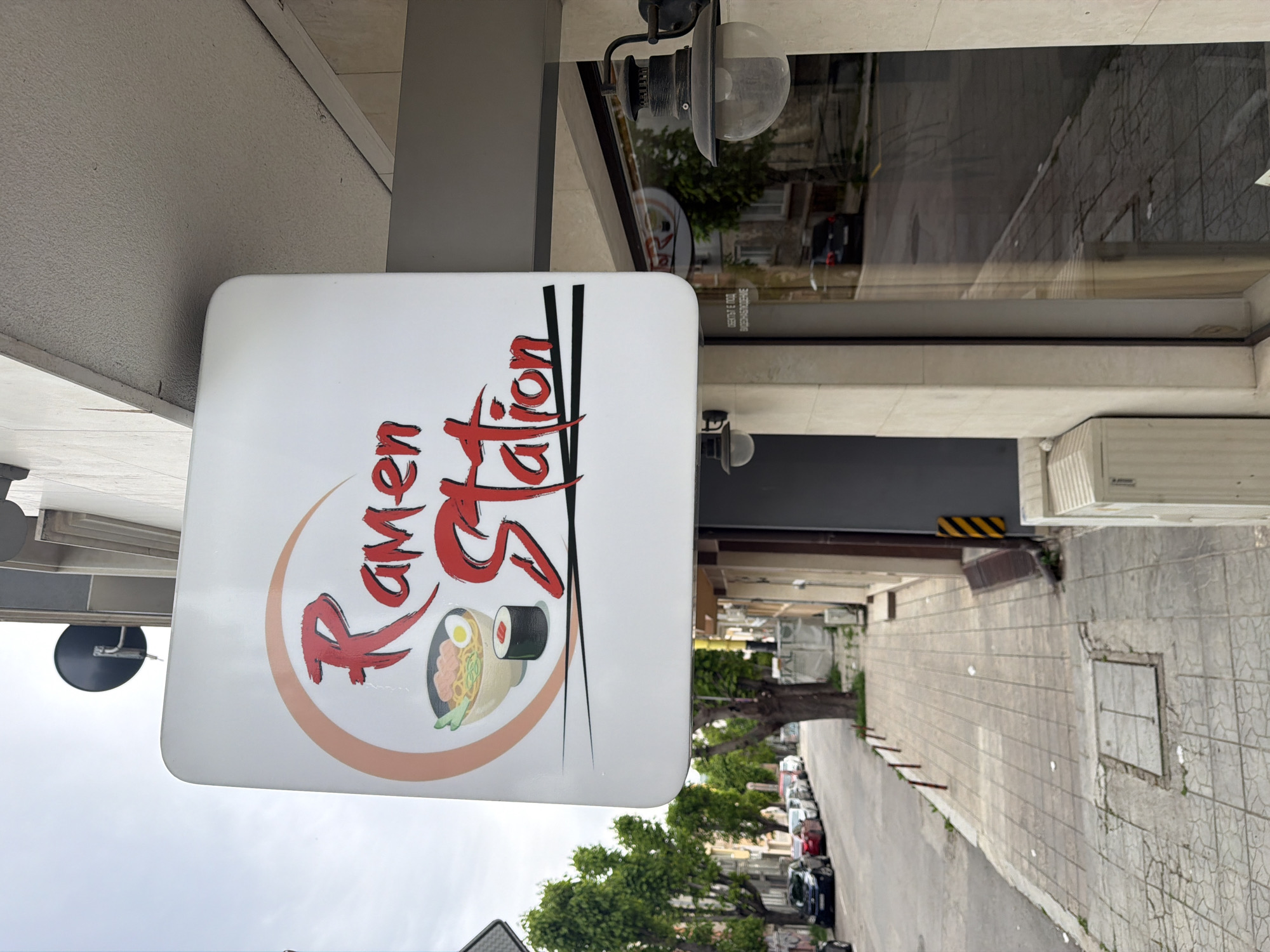 The image shows a square sign that reads "Ramen Station" in stylized red font, featuring a drawing of a bowl of ramen and sushi within an orange circle. The sign is mounted to a building facade with visible light fixtures nearby. The background shows an urban street in Varna, Bulgaria, featuring parked cars, trees, and tiled sidewalks. The building's facade includes windows with reflective surfaces, and there is a glimpse of an air conditioning unit mounted on the wall.