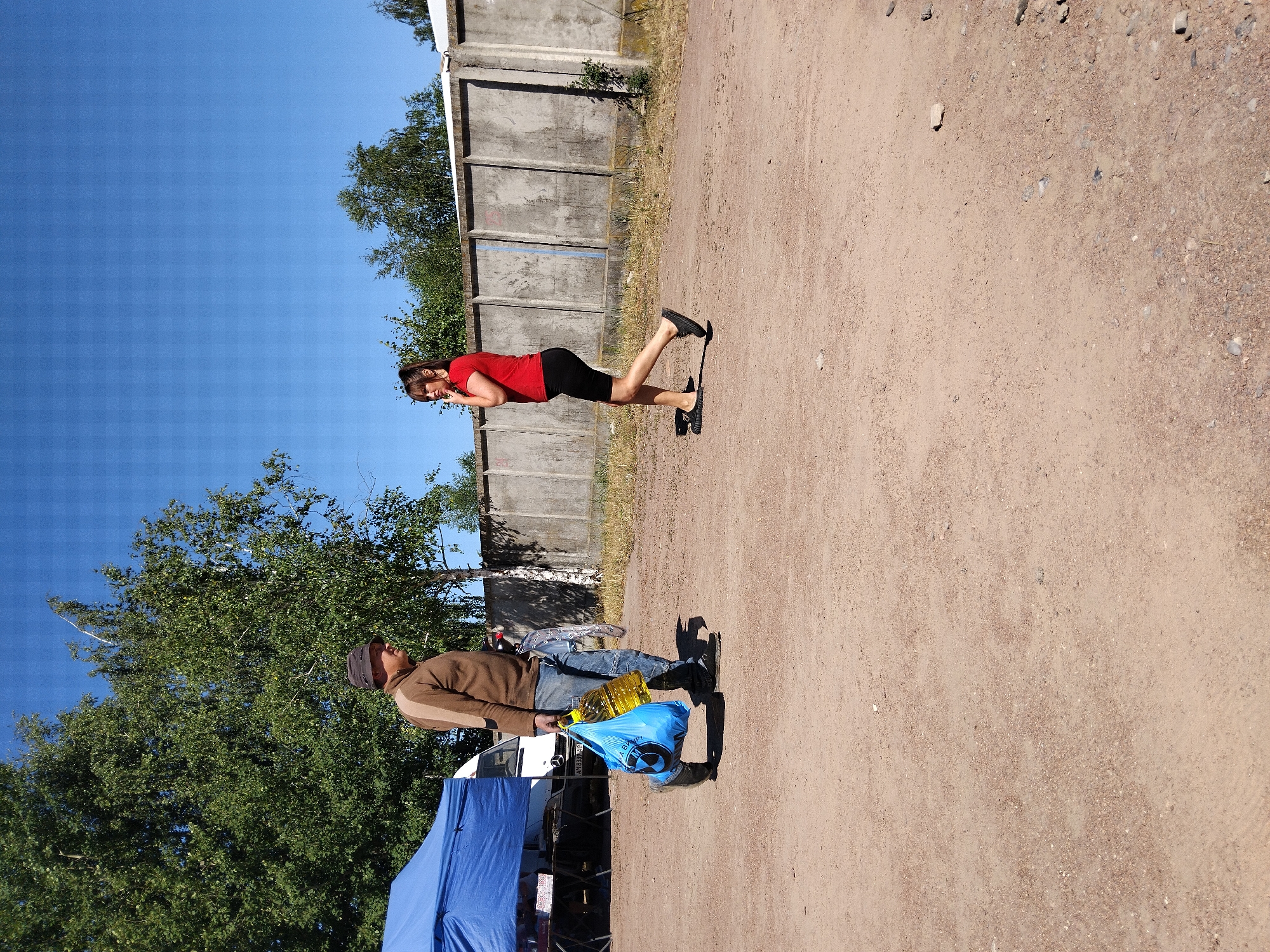 In Irshansk, Ukraine, on a clear, sunny day, a man and a woman are depicted in a sparsely populated outdoor area. The ground is covered in light brown dirt with scattered small stones and debris. In the background, a grey concrete wall runs horizontally across the scene, with trees and foliage visible beyond it.
The man, positioned on the left side of the frame, wears a brown long-sleeved shirt, blue jeans, and a dark hat. He is carrying a large bright blue plastic bag in his left hand, which contains a large yellow plastic bottle. His right hand is partially obscured by the bag. He appears to be walking.
To the right of the man, the woman is walking away from the camera. She is wearing a red short-sleeved t-shirt and black shorts. Her right hand is raised to her face, and she is looking to her left. She is wearing black slip-on shoes.
Further to the left, a blue tent is visible, along with a white vehicle and a folding table, suggesting a possible market or roadside setup. The overall scene conveys an ordinary day outdoors with two individuals in close proximity.