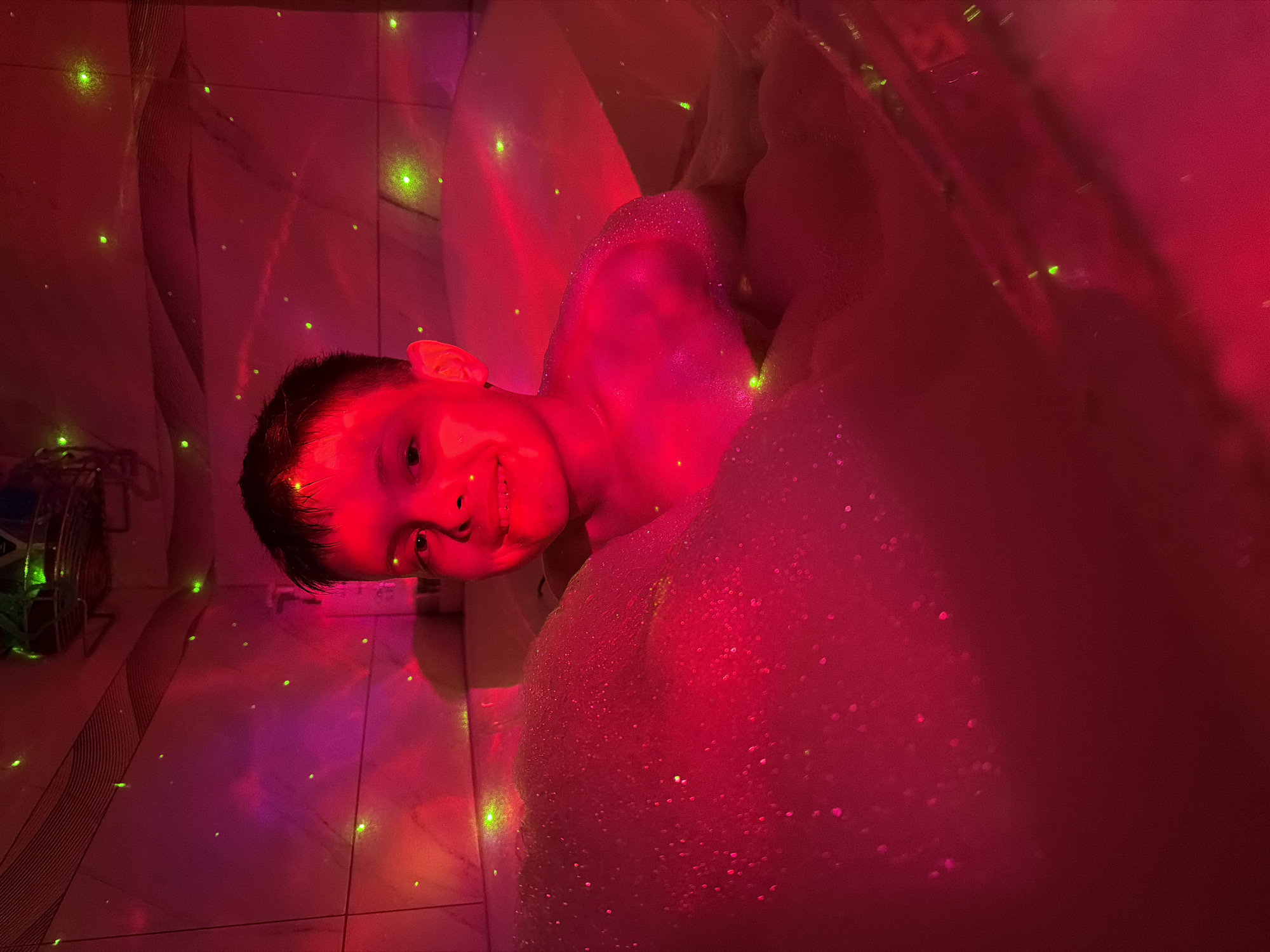 A young boy is visible in a bubble bath, illuminated by a red light and projected green dots. He is partially covered in foam and is smiling at the camera. The scene appears to be indoors, likely in a bathroom due to the tiled floor. There is a metal object with green details on the left side of the image. The location is unknown and cannot be verified from the image.