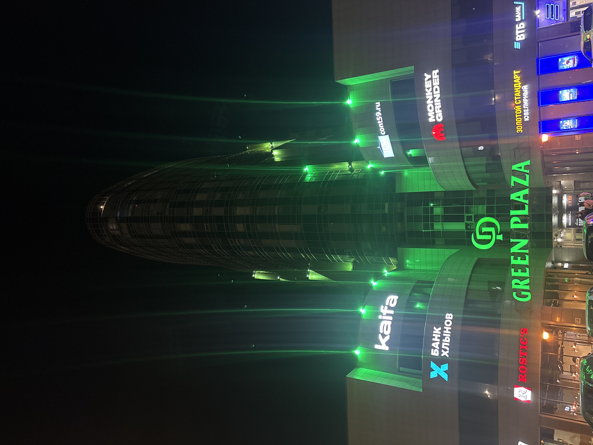 The image captures a night view of the Green Plaza business center in Perm, Russia. The architecture features a rounded, oval-shaped building on the left, with a more standard, block-like structure on the right. The buildings are illuminated with green lighting, creating a prominent visual effect.
Various business logos and signage are visible on the right side of the image, including "Kaifa," "Банк Хлынов," "Rostic's," "Green Plaza," "Monkey Grinder," "Золотой Стандарт Ювелирный," and "ВТБ Банк." The text is a mix of Russian and English.
The image is taken at night, with the surrounding environment appearing dark. The presence of cars and building lights indicates that the location is within an urban area.