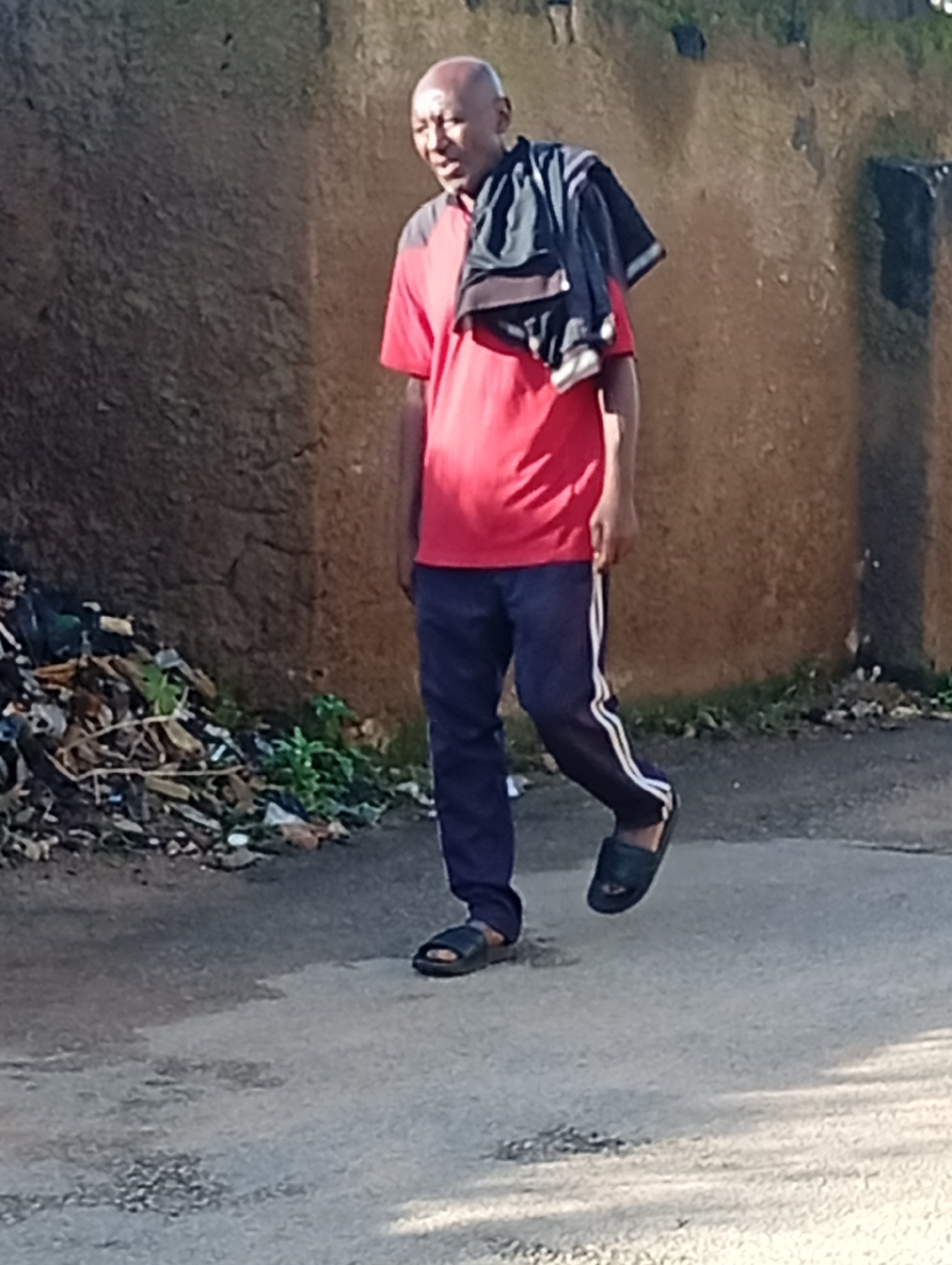 A bald, middle-aged man with dark skin walks on an asphalt road in Gura Top, Nigeria. He is wearing a red t-shirt, dark blue track pants with white stripes, and black sandals. A dark-colored jacket or sweater is draped over his left shoulder. The man is looking to his left, and his mouth is slightly open. The background consists of a rough, textured brown wall and a pile of debris to the left of the man, which includes plastic bags, branches, and other discarded items. The road surface shows signs of wear and has several small patches of damage. The lighting suggests it is daytime, with shadows indicating the direction of the sun.