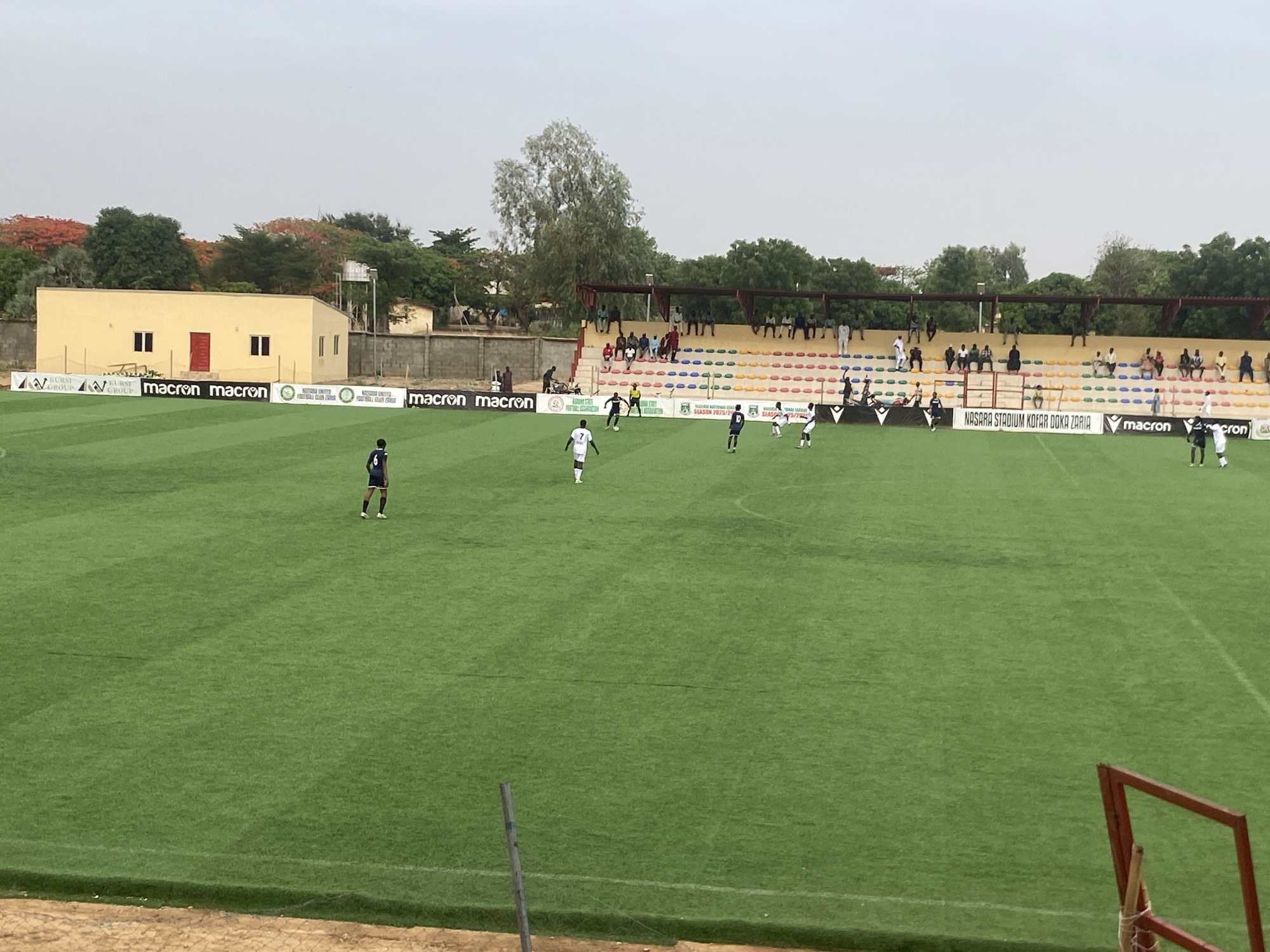 A soccer match is in progress on a well-maintained green field, likely at the Nasara Stadium Kofar Doka Zaria, as indicated by signage. Several players are visible in white and blue uniforms, with one player in a blue uniform standing distinctly on the left side of the field. Spectators are seated in stands located behind the field. A yellow building with a red door is positioned behind the sideline, marked with "macron" logos and other sponsor banners. Trees with green and orange foliage border the stadium. The sky is overcast, creating diffused lighting across the scene. The location is Tudun Wada, Nigeria. A portion of a red-painted metal goalpost is visible in the lower right corner.