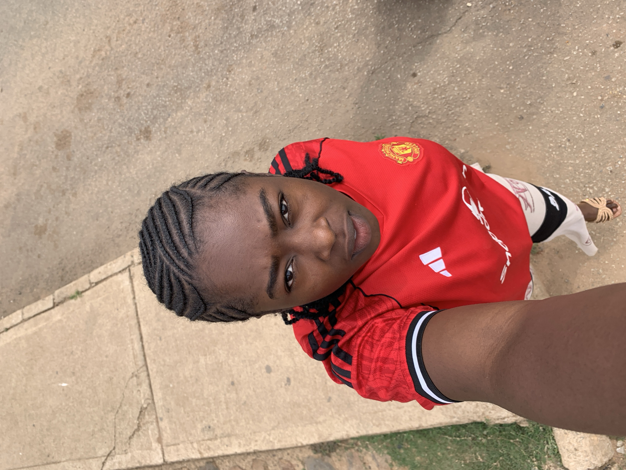 A young person, likely female, with dark skin and braided hair, looks directly up at the camera in what appears to be a high-angle selfie. She is wearing a red short-sleeved sports jersey with black and white striped cuffs. The jersey features a yellow circular "MANCHESTER UNITED" logo with a ship and devil emblem on the left chest, a white Adidas logo on the right chest, and "DXC" in white text on the front. She wears white shorts or trousers with black and white vertical stripes on the sides, and light-colored, possibly gold, patterned sandals on her feet.
The setting is outdoors, with the person standing on a paved surface, likely concrete, which shows varying textures, stains, and cracks. Small patches of green grass are visible along the edges of the paved area. The lighting suggests it is daytime. The image captures a moment of posing for the camera in Rafin Par, Nigeria.