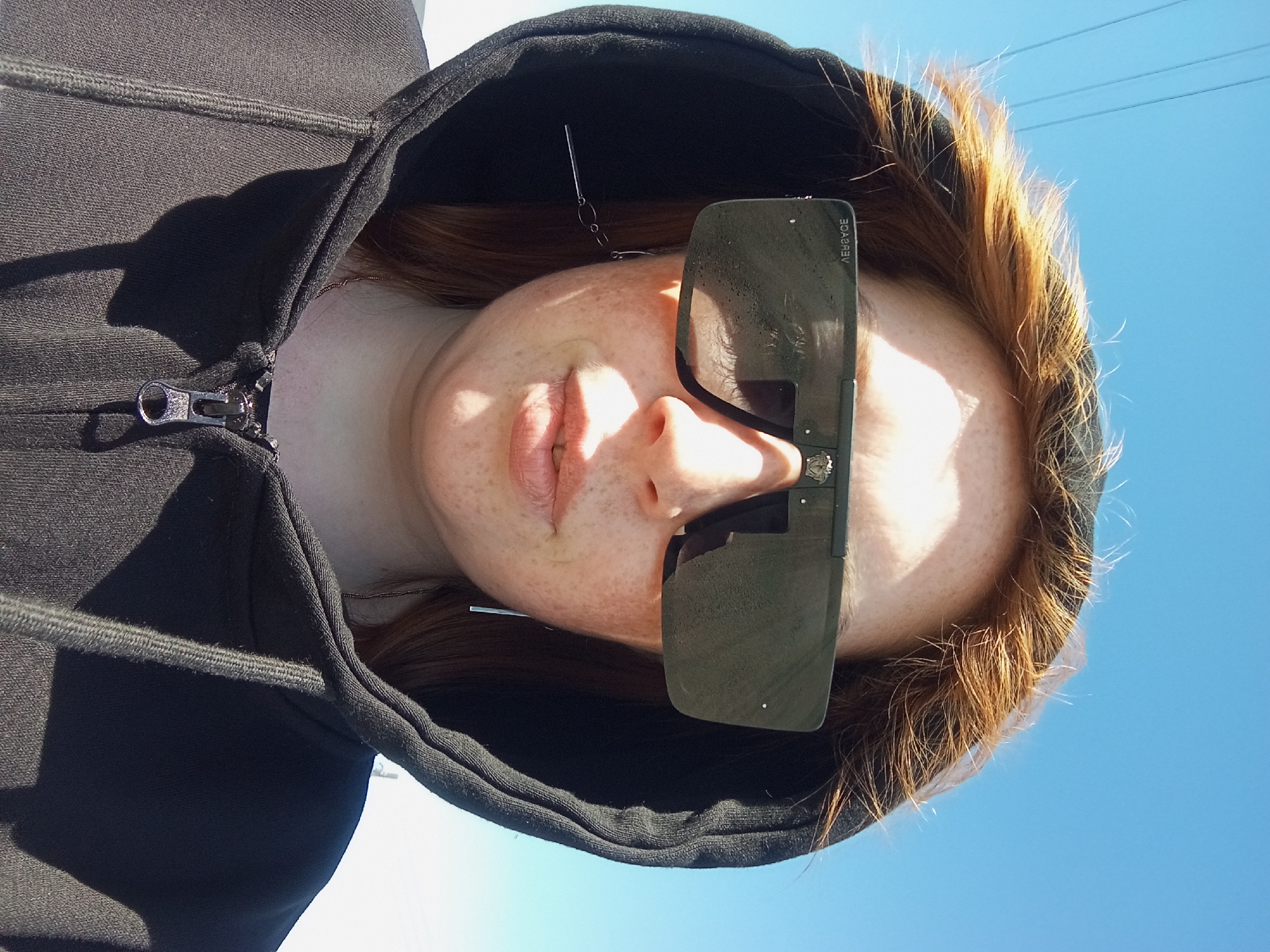 A woman with red hair and freckles is looking down at the camera while wearing a black hoodie and large sunglasses. The background is a bright blue sky, suggesting it is daytime. The photograph appears to be taken outdoors. The location of Yurgamysh, Russia is not verifiable from the image.