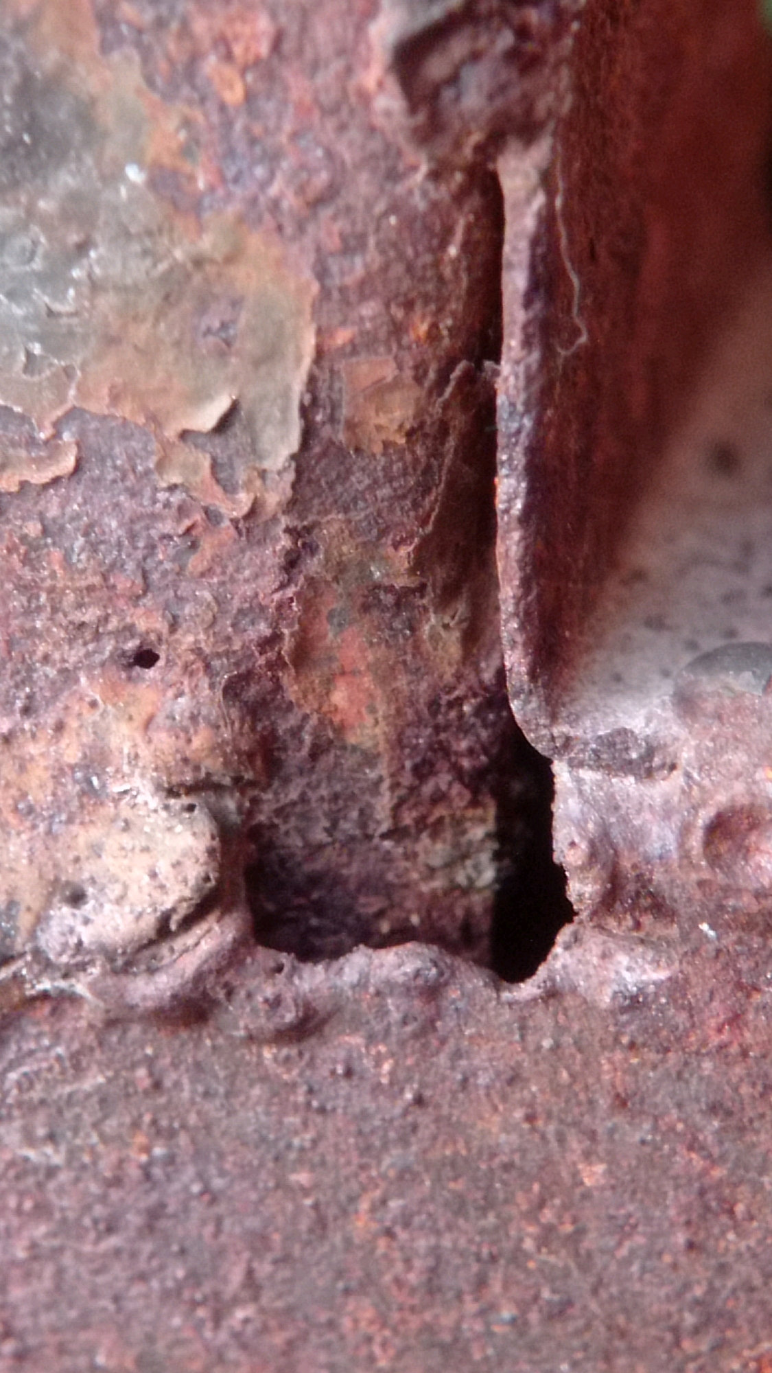 The image displays a close-up view of heavily rusted metal. The texture is rough and flaky, with various shades of brown and red indicating significant corrosion. There is a visible gap or crevice in the metal, revealing darker, more deeply corroded areas within. The surface is uneven and pitted. The location in Mulyoagung, Indonesia, suggests this is likely an outdoor scene, possibly of an aged piece of machinery, infrastructure, or discarded metal in a rural or industrial area. No people, animals, or specific actions are depicted.