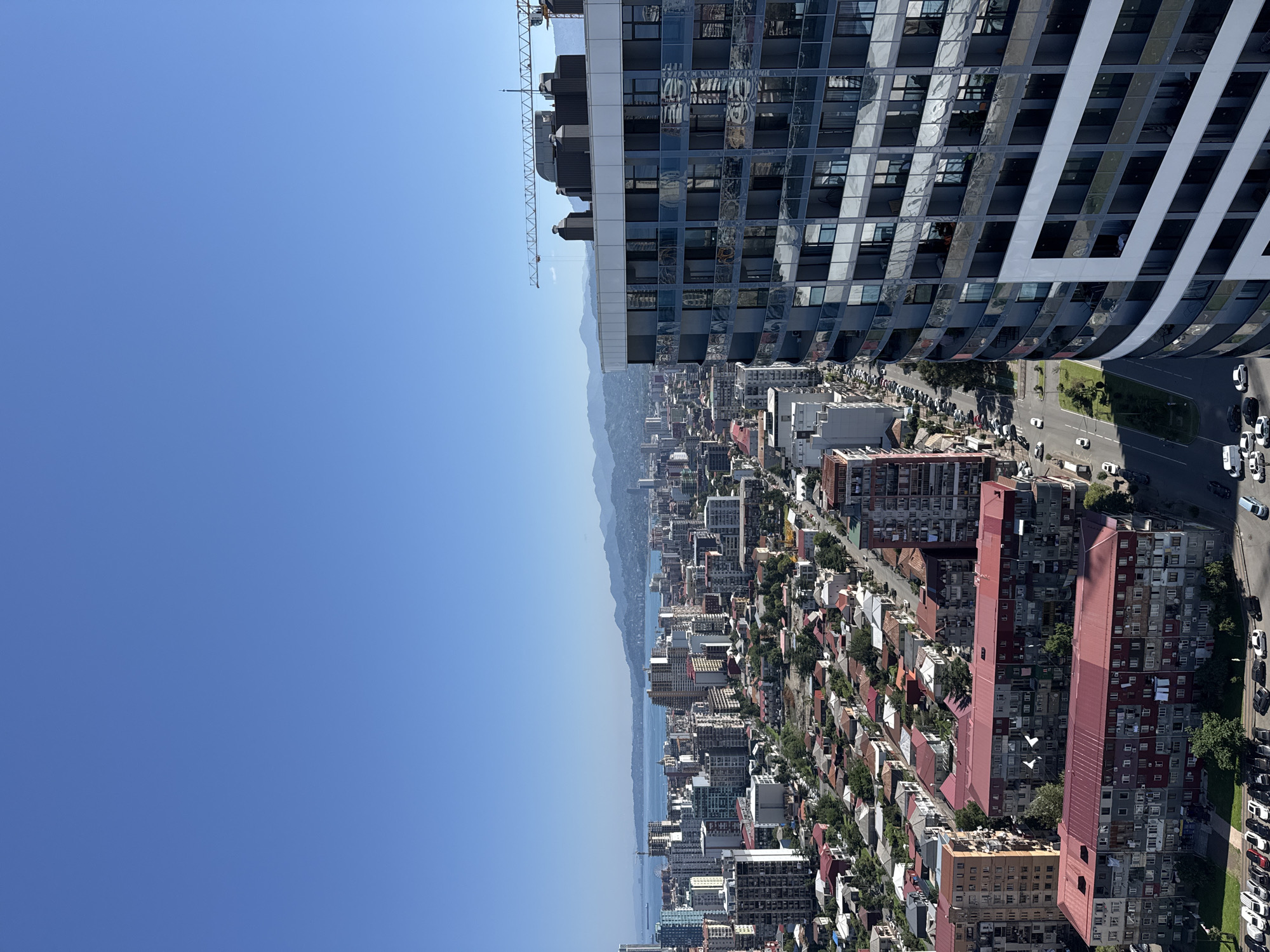 This is a high-angle, wide-angle, outdoor, daylight, vertical photograph of the cityscape of Batumi, Georgia, as viewed from a tall building. The sky is clear and blue, occupying the top portion of the frame. In the mid-ground and background, a dense urban environment unfolds, characterized by numerous multi-story buildings of varying architectural styles and heights. Many buildings are constructed with concrete, and some feature distinctive red roofs. A body of water, likely the Black Sea, is visible in the distance, with rolling, verdant hills or mountains forming the horizon line.
In the foreground, to the right, a modern, curved skyscraper with a reflective glass facade dominates the frame. This building appears to be under construction, as evidenced by a construction crane positioned on its roof. The glass panels reflect the surrounding environment and sky.
Below the buildings in the mid-ground, a network of streets is visible, with cars and a bus navigating the roads. Small residential buildings with red roofs are clustered in the lower mid-ground, interspersed with trees and green spaces. A parking lot filled with cars is present in the lower left portion of the image.
The overall impression is a vibrant, growing city with a mix of older and newer construction, set against a backdrop of natural beauty. There are no discernible people or animals in the photograph. The scene depicts a typical day in a coastal urban environment.