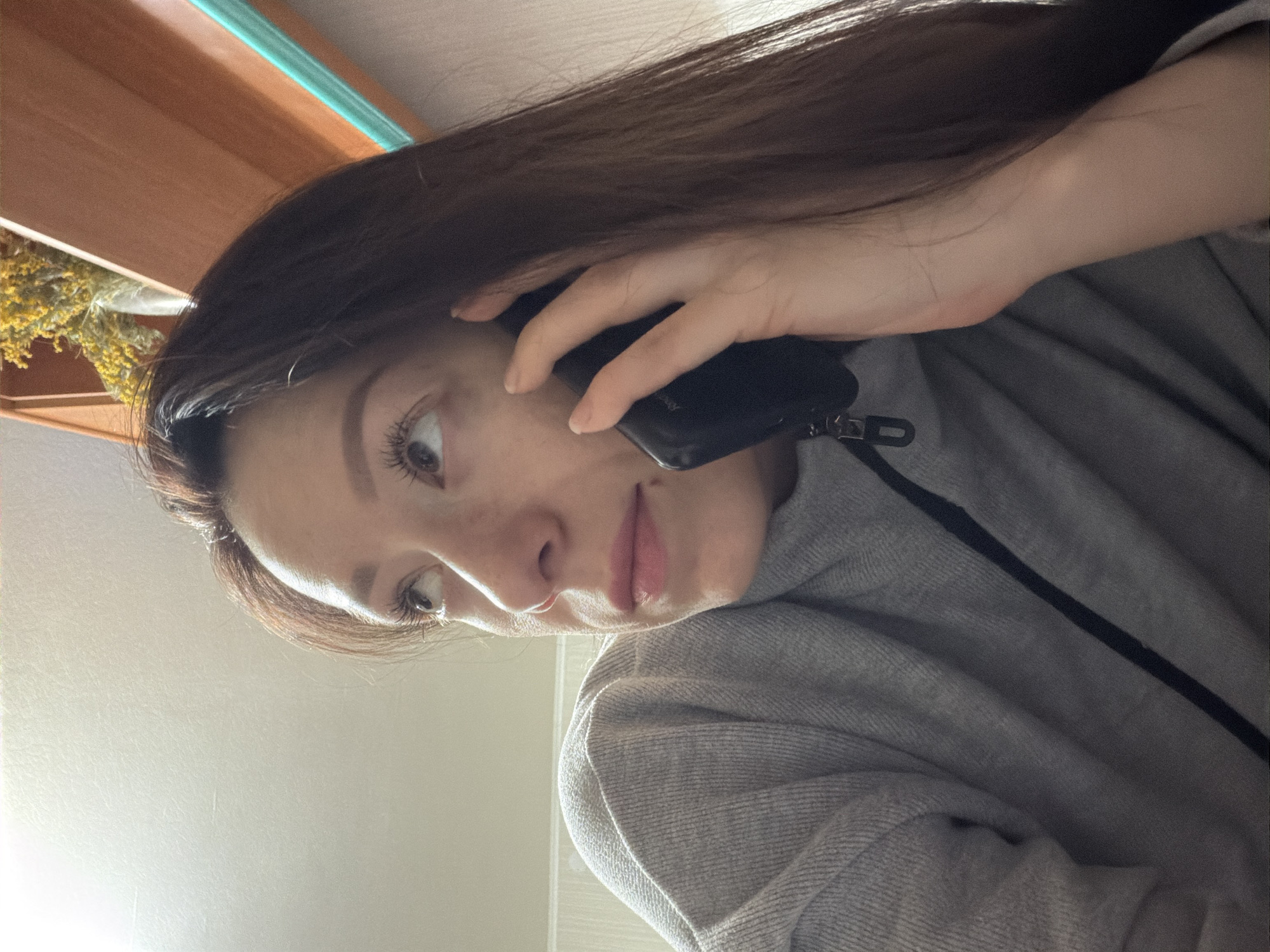 A woman with long brown hair is holding a black mobile phone to her left ear. She is wearing a gray sweatshirt. The background is a room with wooden furniture. There is also a plant in the background. The location is Unknown, Unknown.