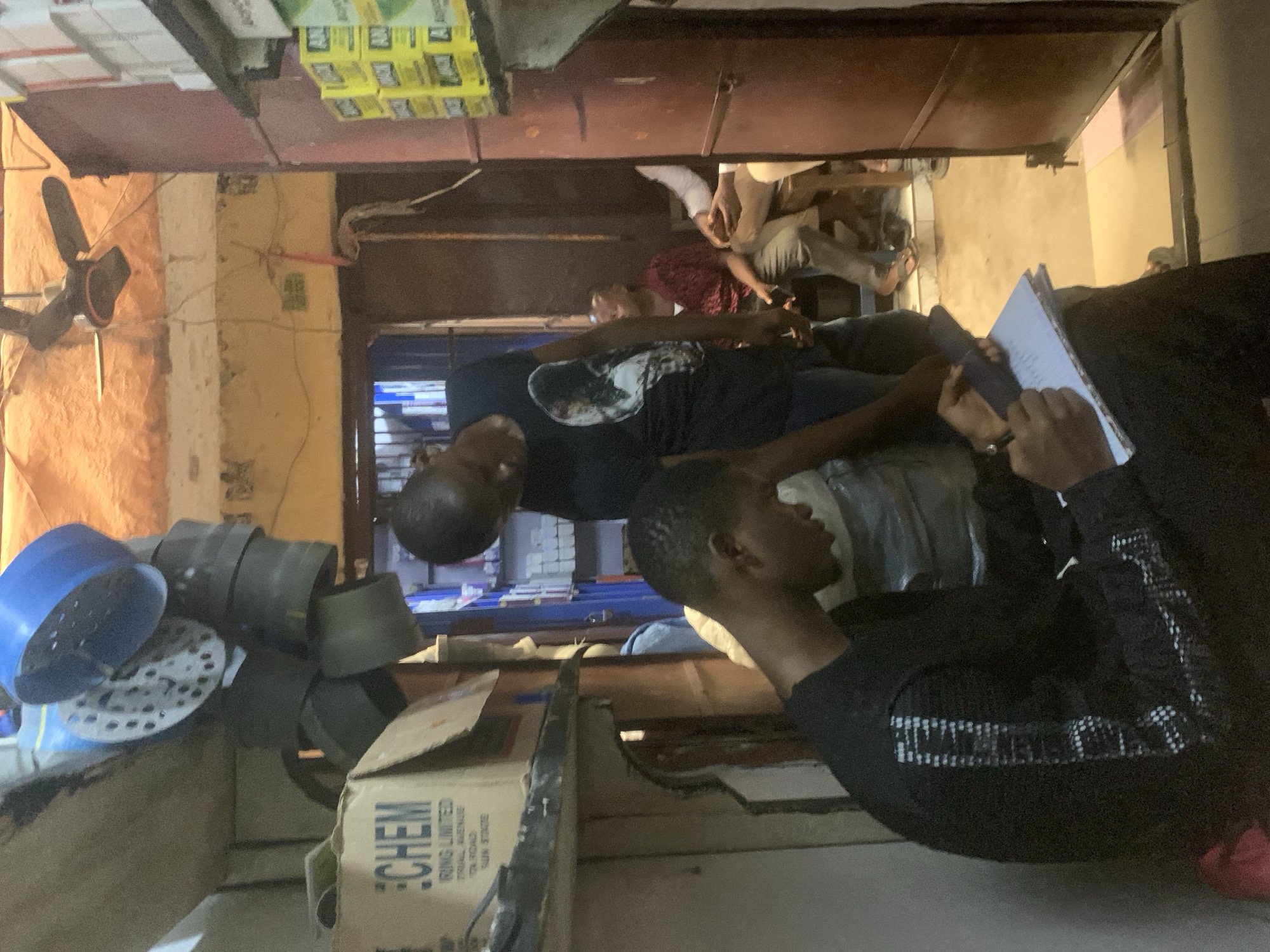 In Fegge, Nigeria, an indoor scene shows two men, one in a black shirt writing in a notebook, and another wearing a black t-shirt with a design, looking at the notebook. Other individuals are in the background. The environment includes boxes, including one labeled "CHEM", and various objects such as blue and grey containers, and a fan.