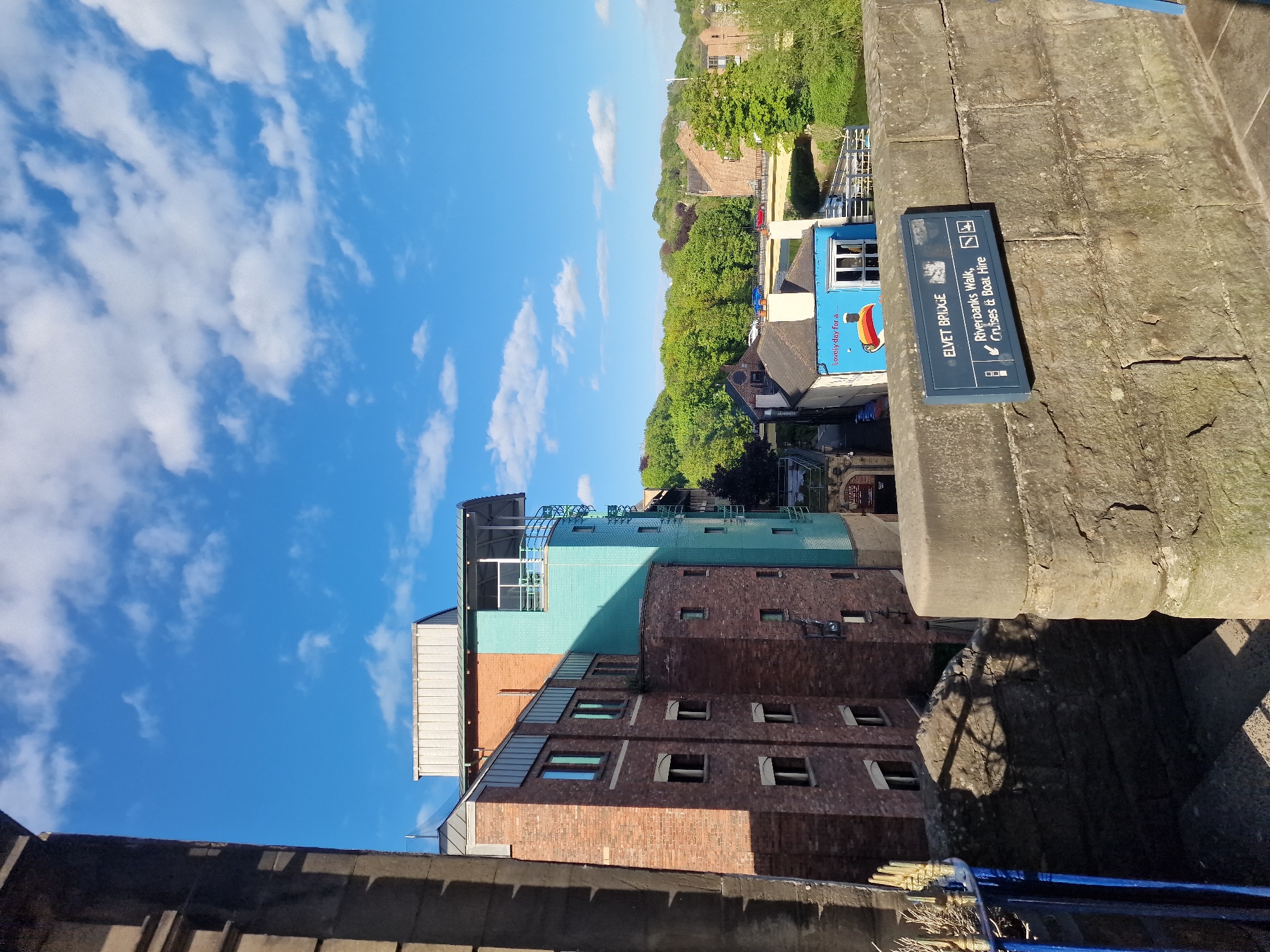 The image is a ground-level view of the Elvet Bridge in Durham, United Kingdom. The sky is blue with scattered white clouds. The bridge's stone structure occupies the right side of the image, with a sign attached to it that reads "ELVET BRIDGE," then lists options for "Riverbanks Walk, Cruises & Boat Hire." A building of varied architectural styles is visible below. It is primarily red brick with several rectangular windows, with additional sections that are teal, light gray, and copper. Adjacent to the building is a view of trees and a building that has a sign that states: "Lovely day for a..." with an umbrella image. A blue railing is visible in the lower foreground.