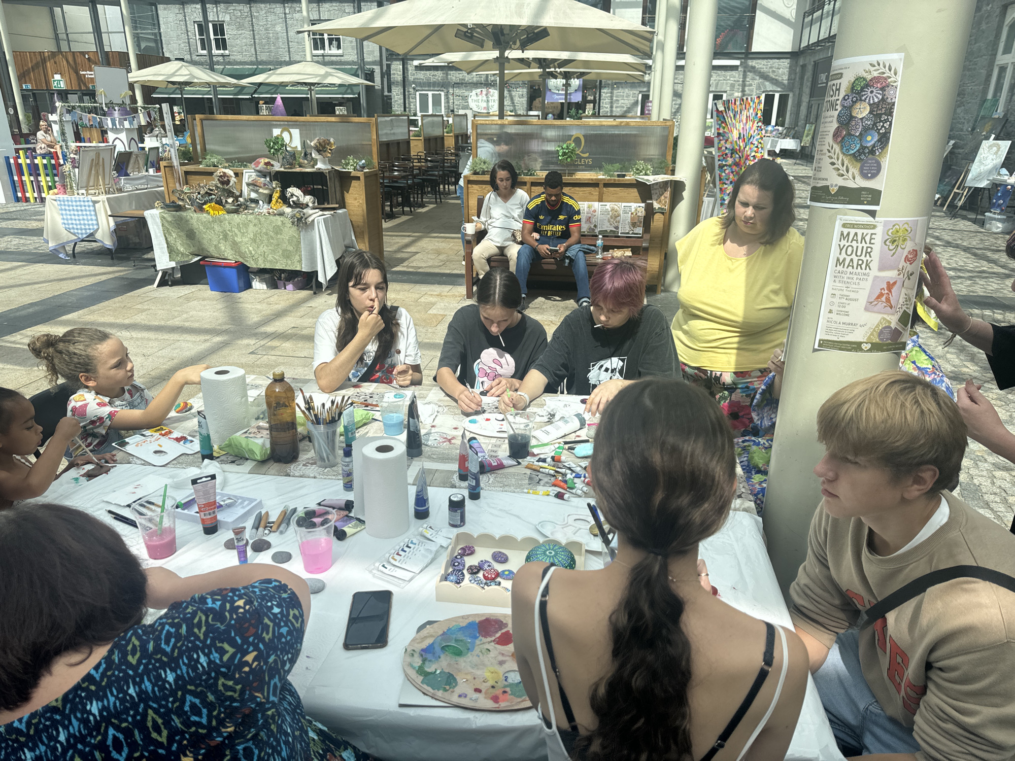 A group of people are engaged in a painting activity at an outdoor event in Kilkenny, Ireland. In the foreground, a long table is set up with art supplies, including paint tubes, brushes, water cups, and palettes. Several individuals, primarily young people and children, are seated around the table, actively painting on rocks and canvas. One young girl with curly hair and a white top is painting with a brush, while another child with a lollipop is focusing on their artwork. A woman in a yellow shirt is standing to the right, observing the activity. In the background, there are market stalls displaying various items, and people are milling about. The overall scene suggests a community art event or workshop taking place in an urban outdoor setting, possibly in a public square or market area of Kilkenny.
