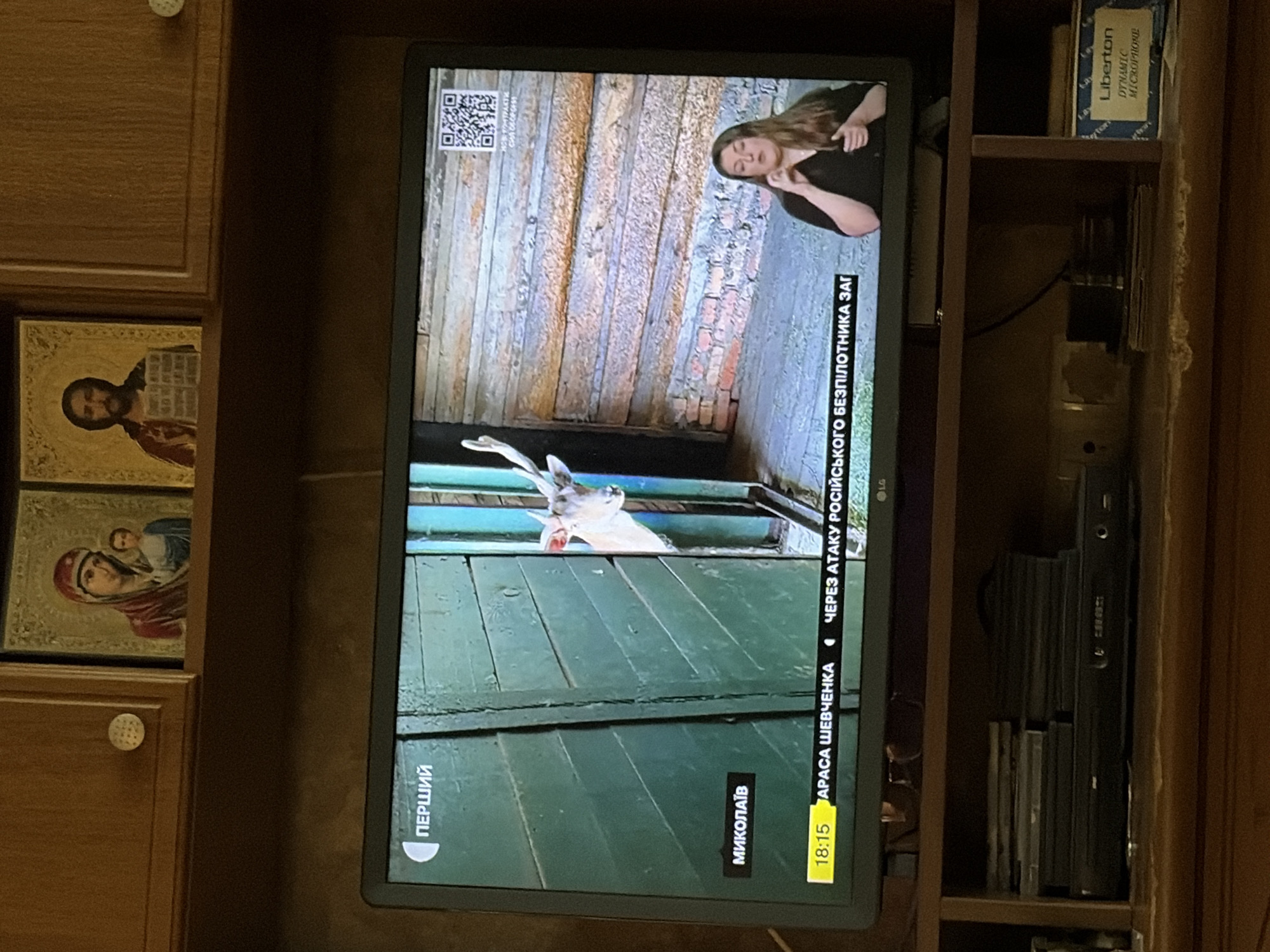 On a television screen, a news report is being broadcast from "ПЕРШИЙ" channel. The screen displays a segment about "МИКОЛАЇВ" and a report titled "18:15 АРАСА ШЕВЧЕНКА ЧЕРЕЗ АТАКУ РОСІЙСЬКОГО БЕЗПІЛОТНИКА ЗАГ". The visual content of the news report shows a white deer peeking from behind a green wooden door and a woman speaking, likely a news anchor or correspondent, with a QR code visible in the background. The television is placed on a wooden shelf, which also holds a DVD player and a stack of DVDs or cases. Above the television, there are two religious icons on a wooden cabinet. The scene is captured in a dimly lit room.